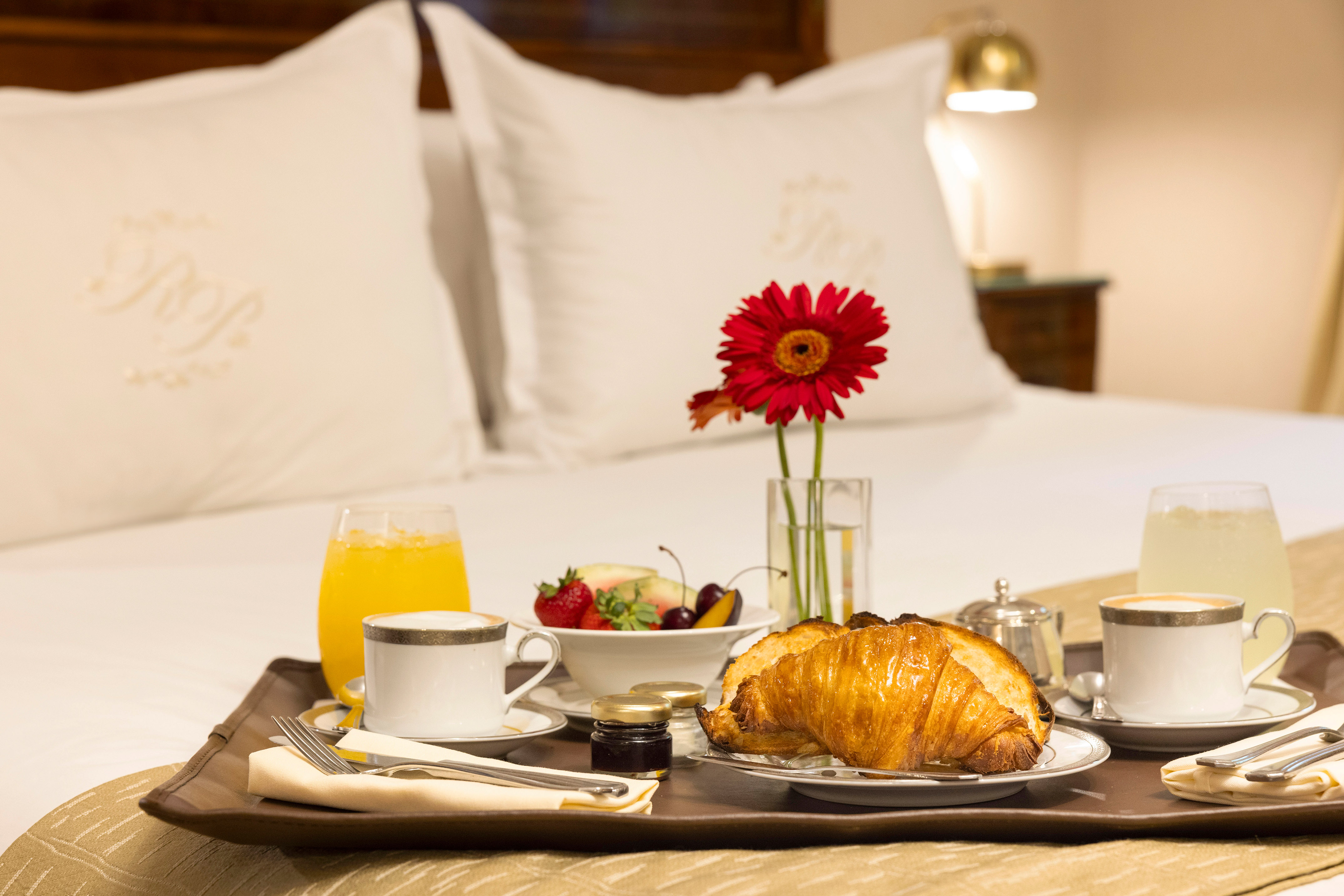 Luxury breakfast in bed: croissant, coffee, juice, fruit, and red flower.