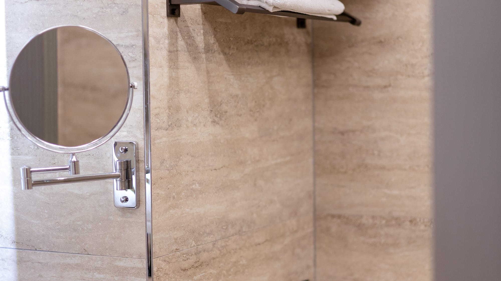 Beige tiled bathroom with white towels on dark shelves and a magnifying mirror.