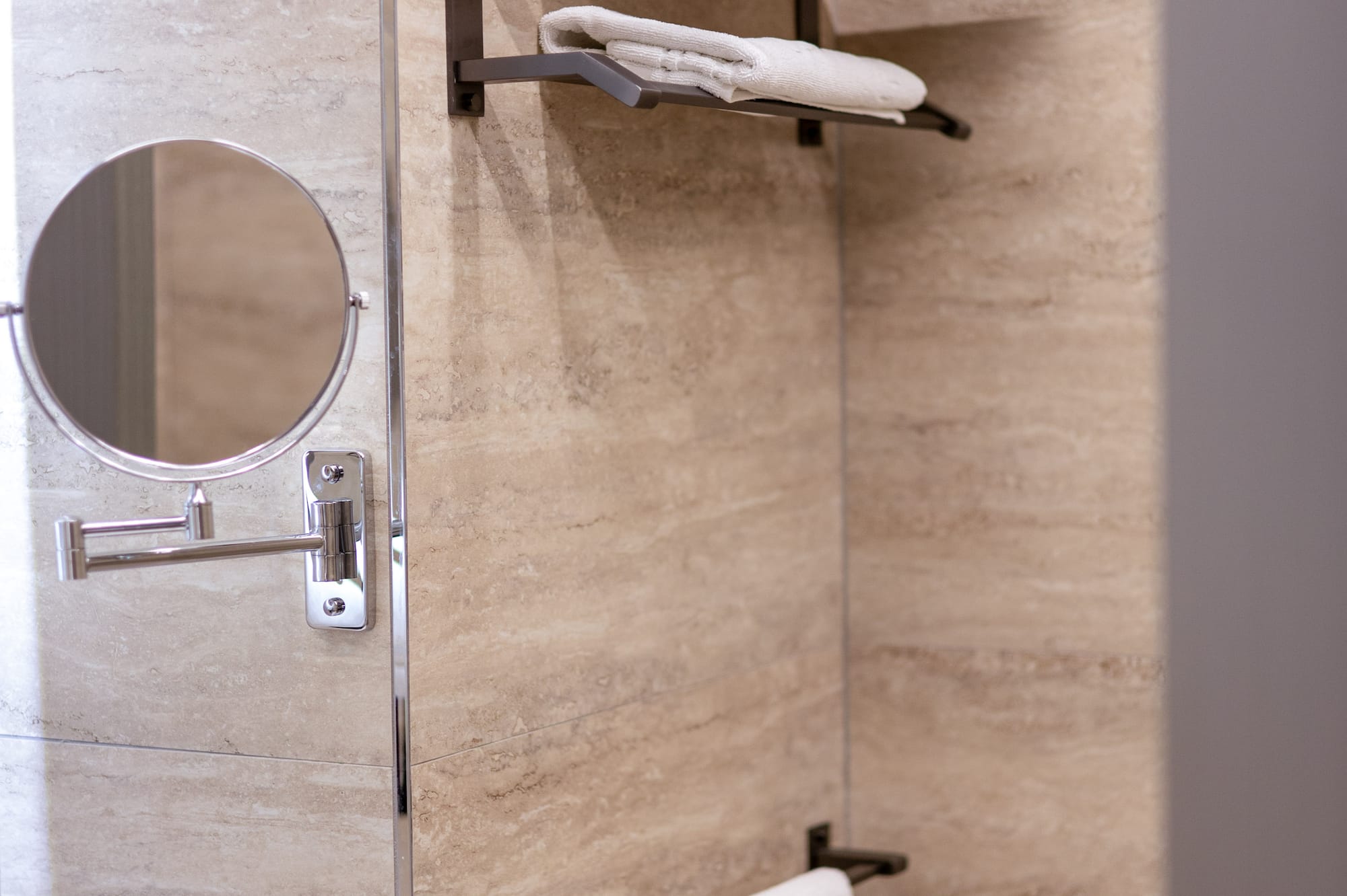 Beige tiled bathroom with white towels on dark shelves and a magnifying mirror.