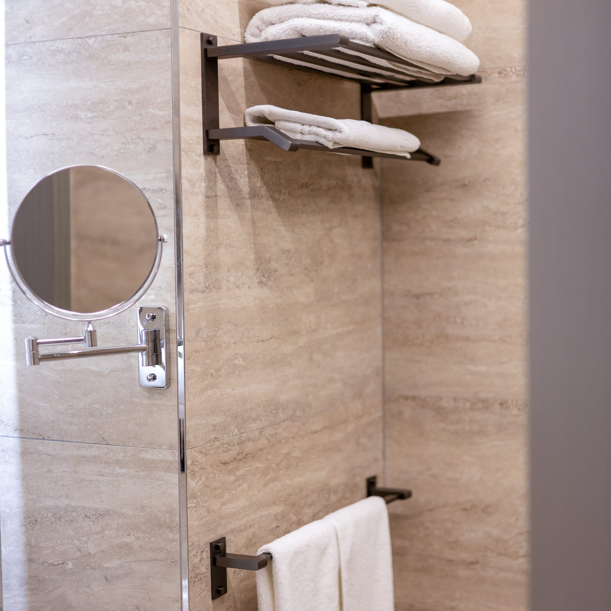 Beige tiled bathroom with white towels on dark shelves and a magnifying mirror.
