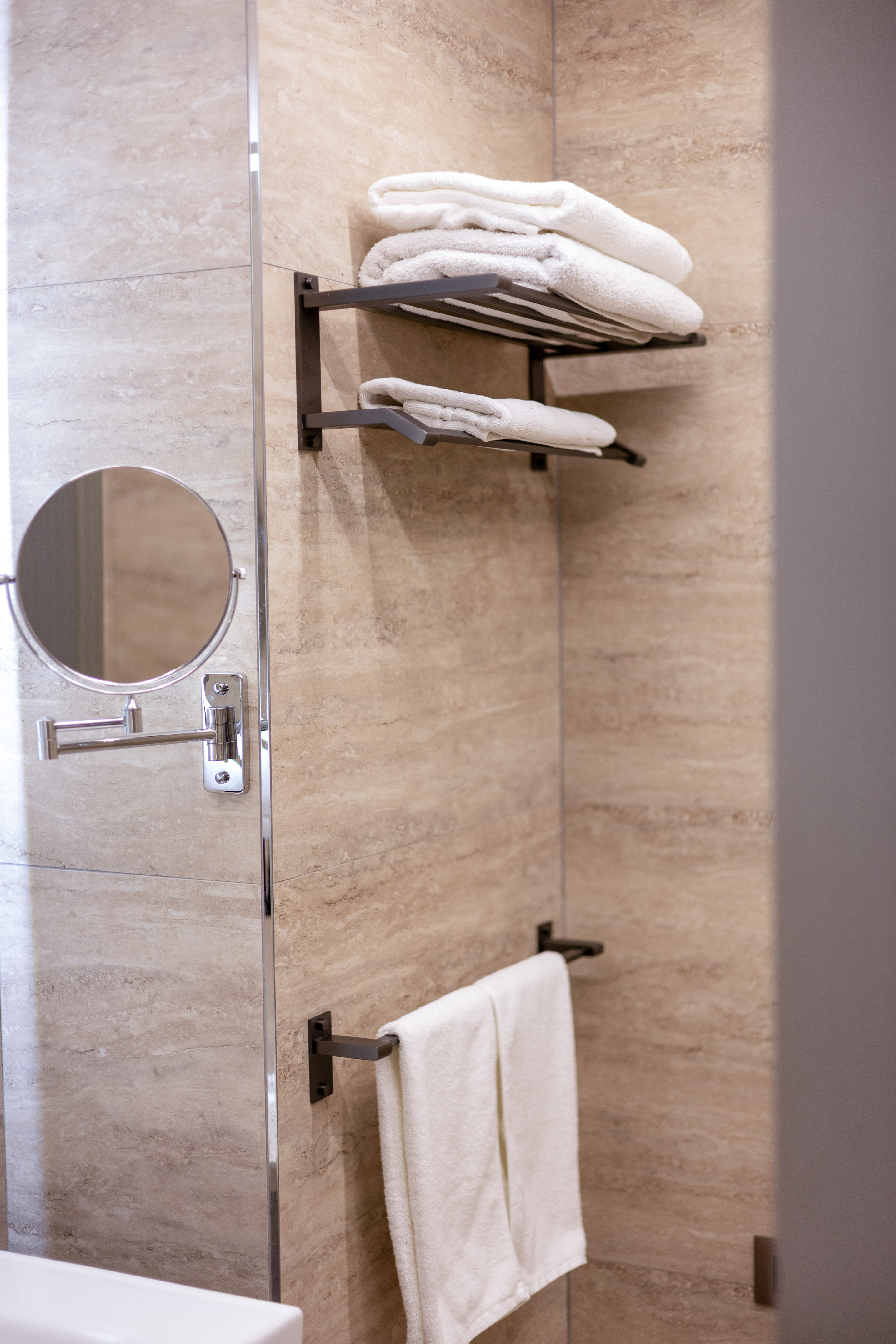 Beige tiled bathroom with white towels on dark shelves and a magnifying mirror.