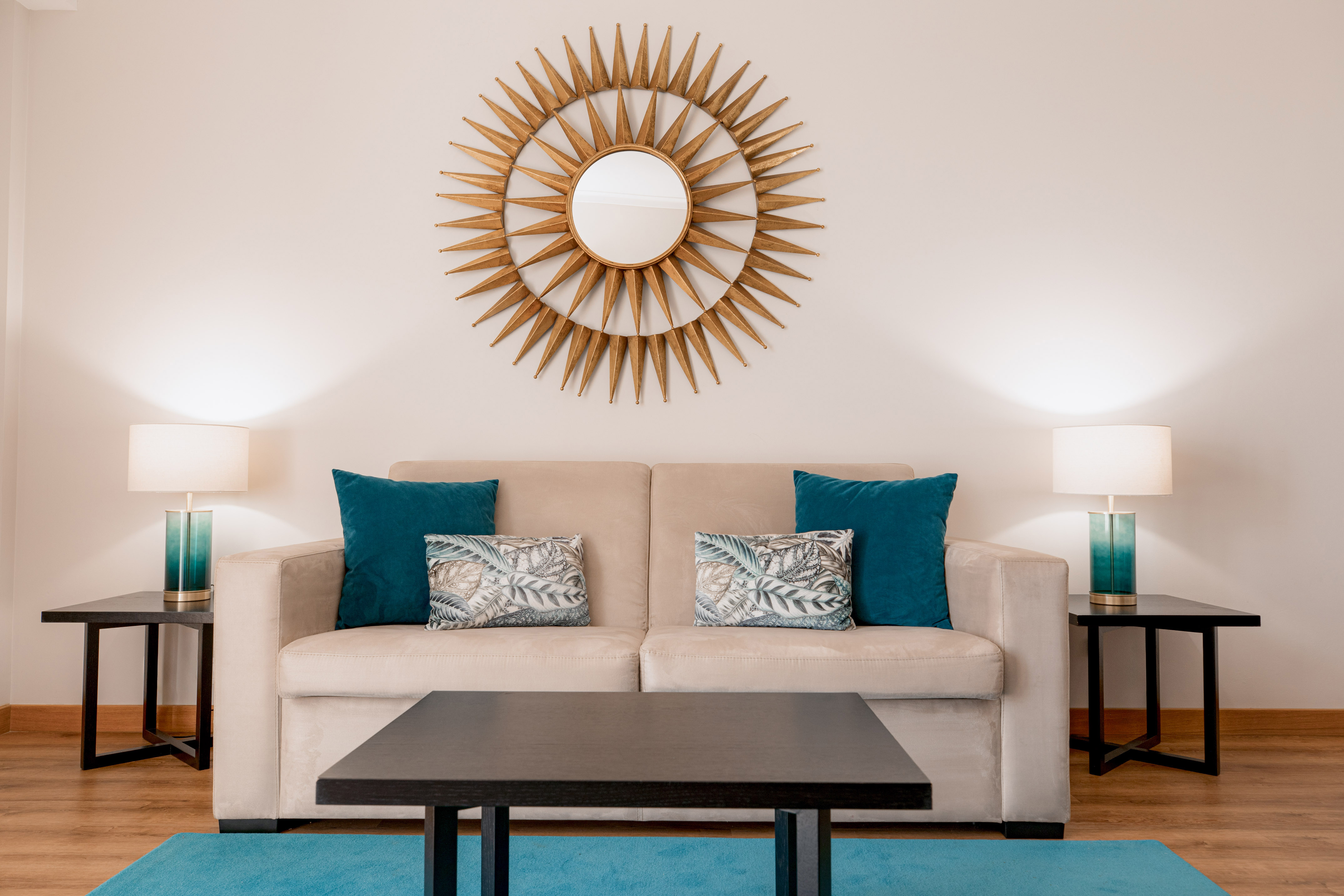 Modern living room with beige sofa, teal accents, and gold sunburst mirror.