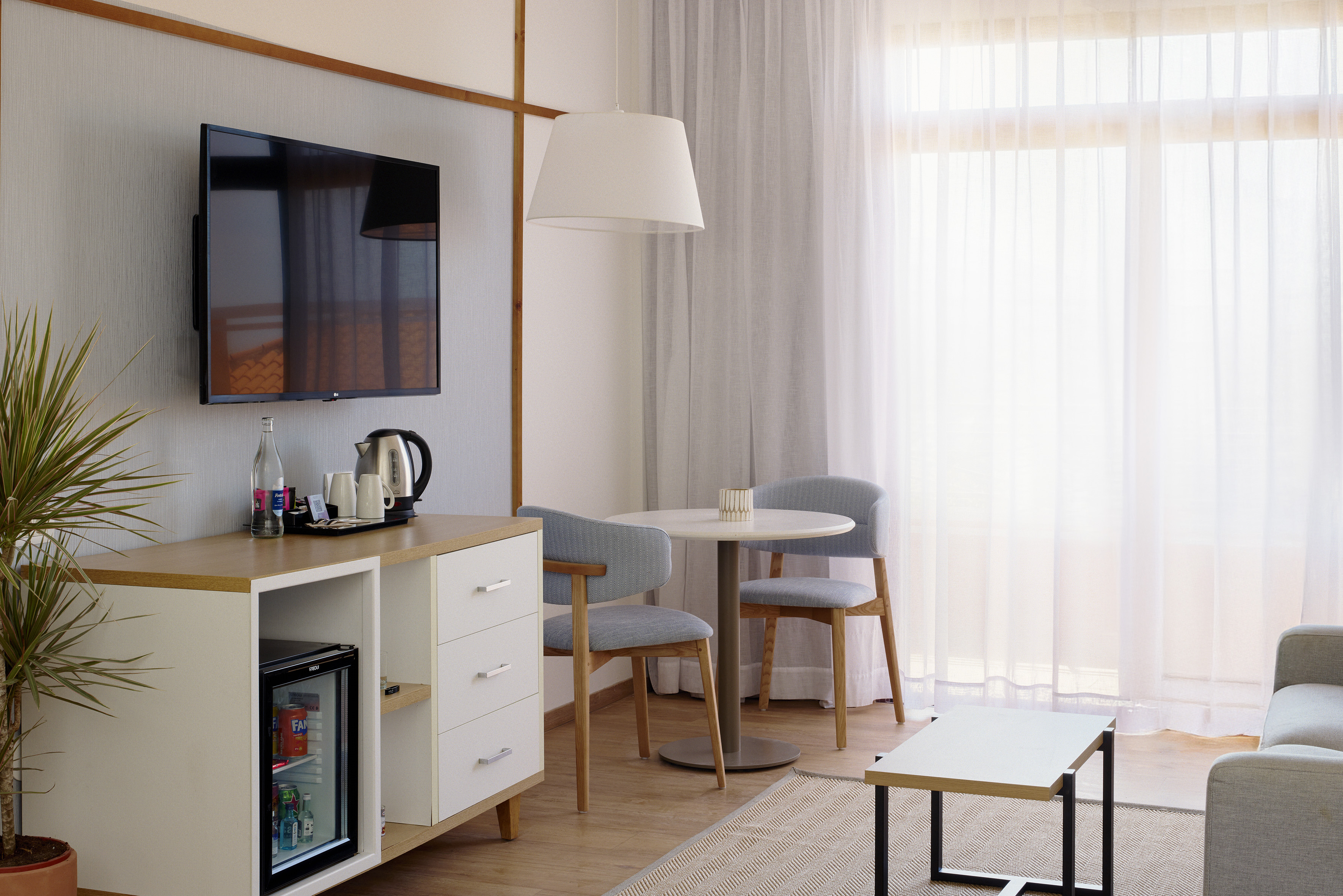 Modern hotel living area with TV, mini-fridge, dining table, and sheer curtains.