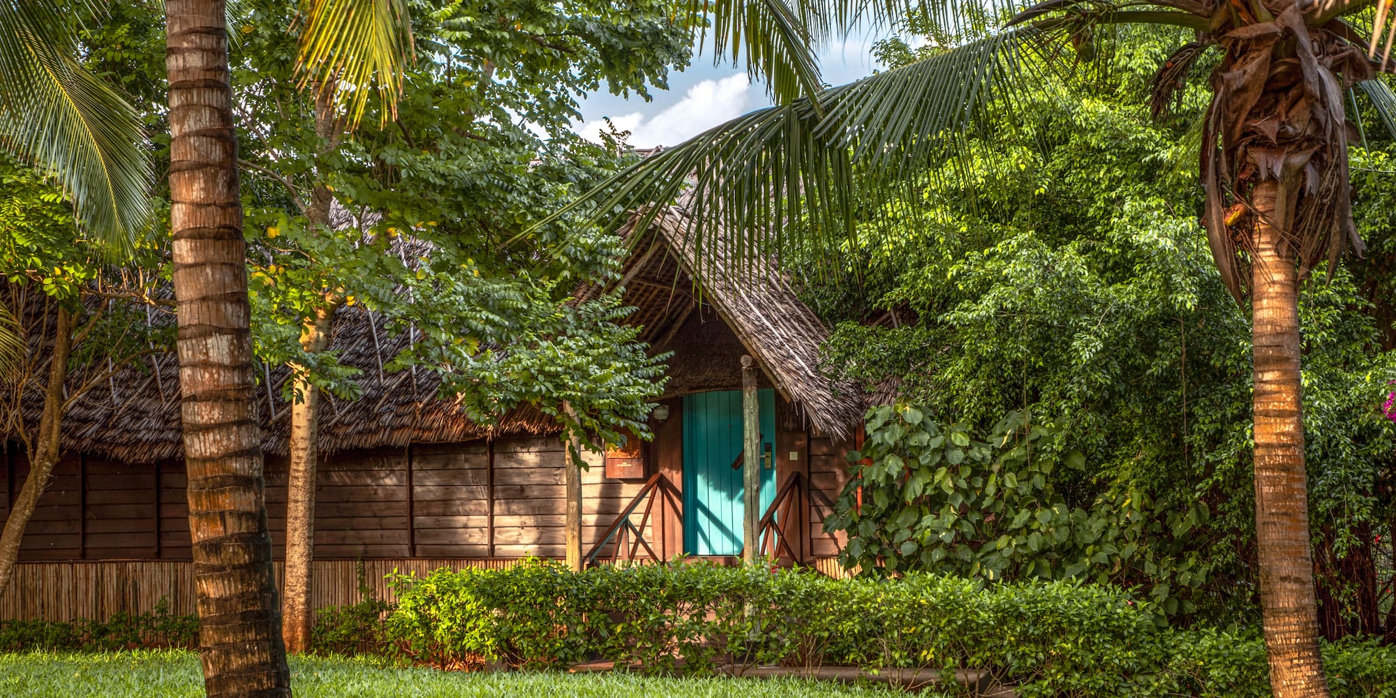 Tropical thatched bungalow with teal door, lush green garden, and palm trees.