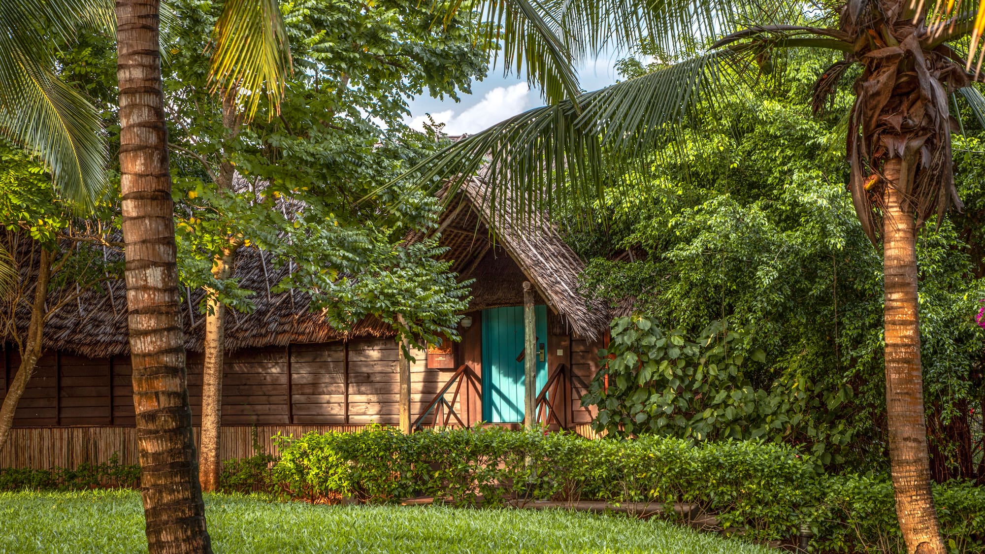 Tropical thatched bungalow with teal door, lush green garden, and palm trees.