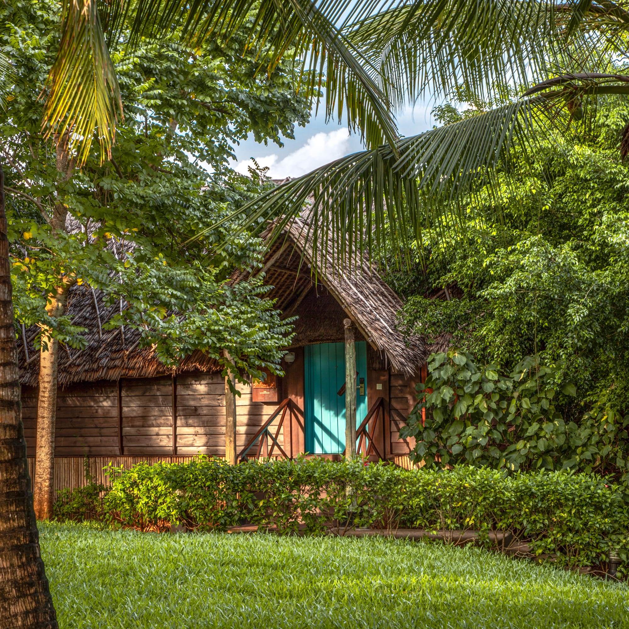 Tropical thatched bungalow with teal door, lush green garden, and palm trees.