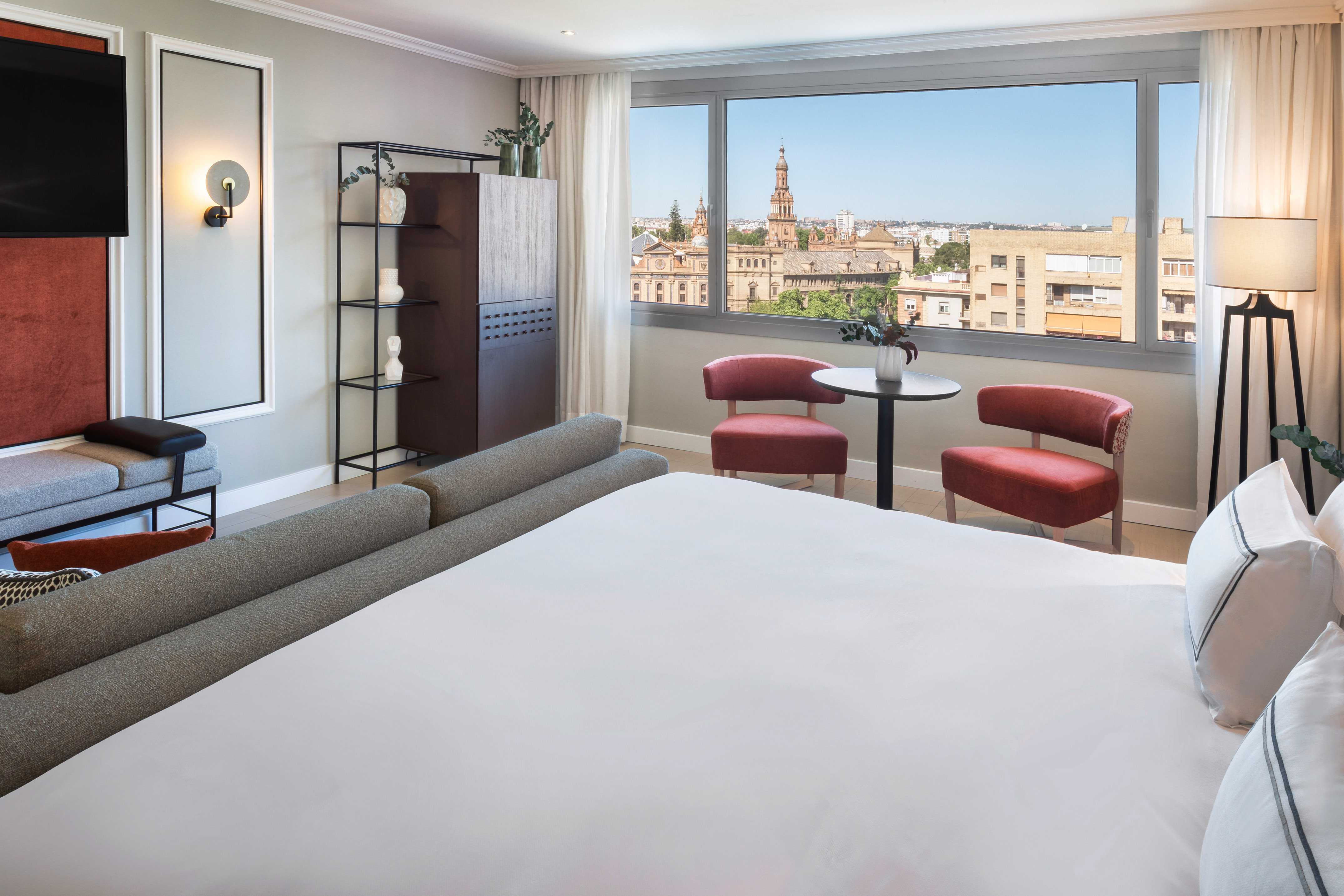 Modern hotel suite with Seville city view, king bed, and stylish red chairs.