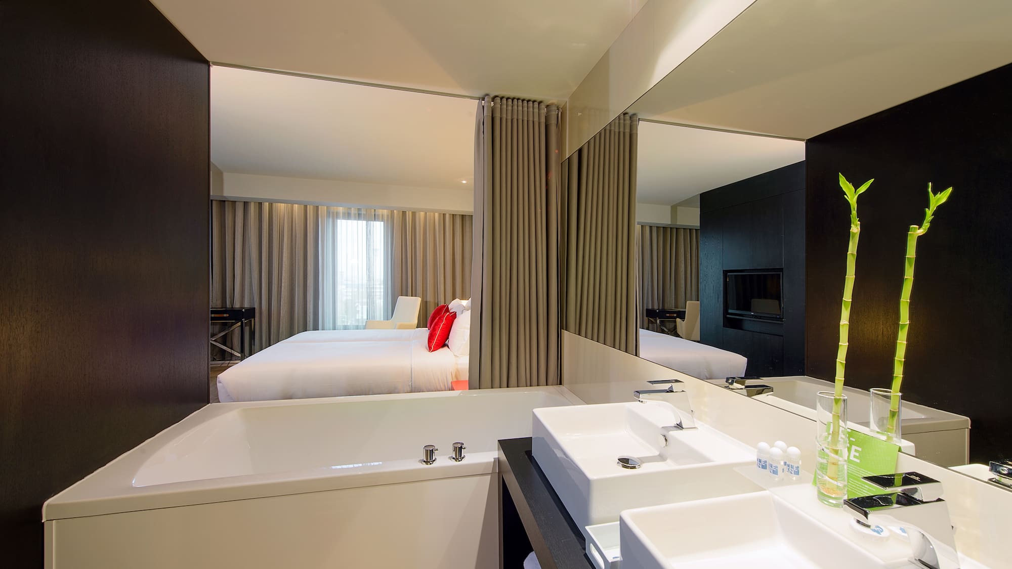 Modern hotel suite bathroom with bathtub, dual sinks, and bedroom reflected.