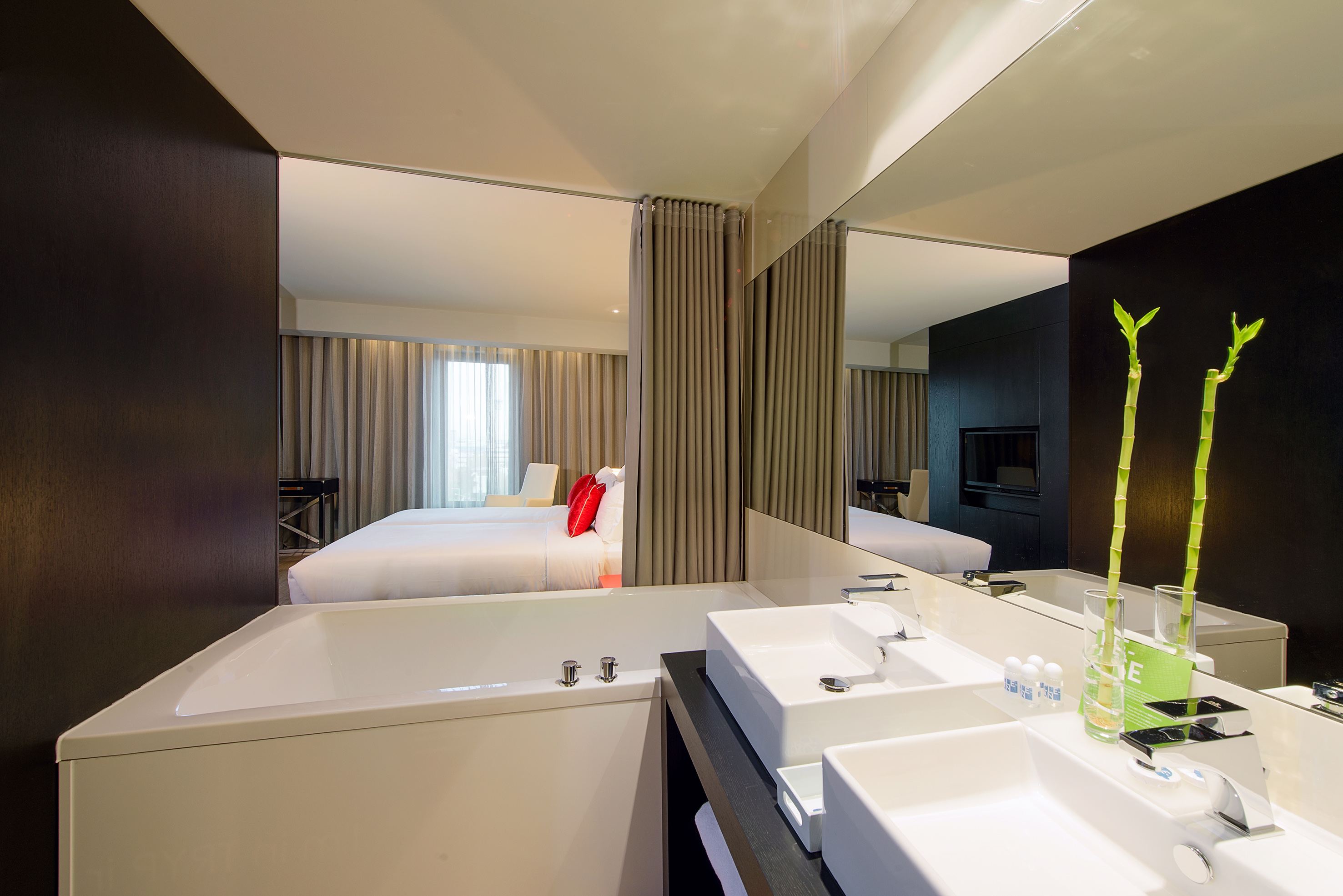 Modern hotel suite bathroom with bathtub, dual sinks, and bedroom reflected.