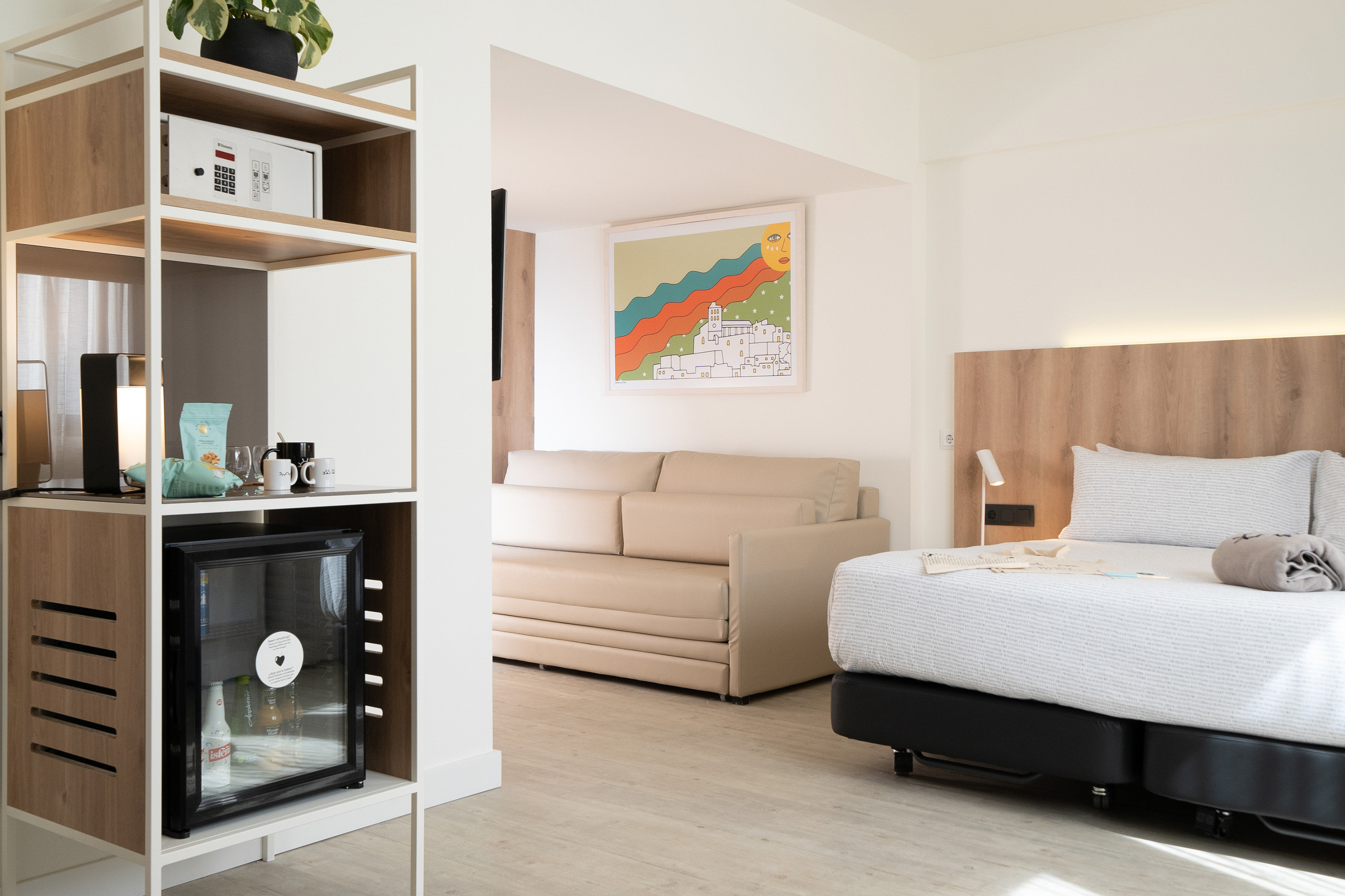 Modern hotel room with bed, sofa, mini-bar, safe, and vibrant artwork.
