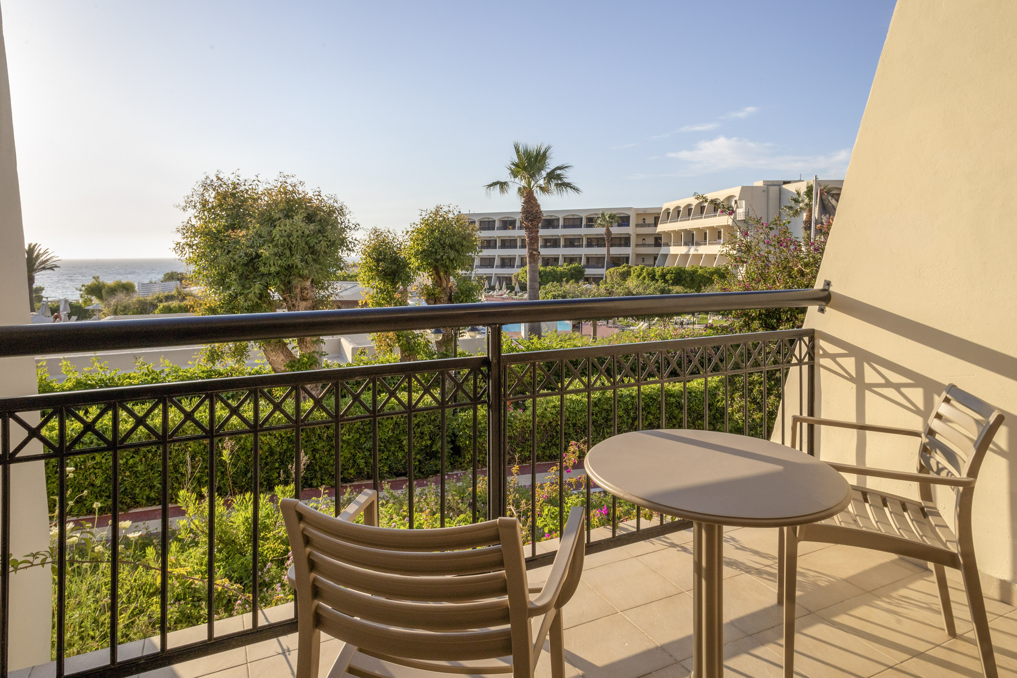 Sunny resort balcony with chairs, table, lush trees, pool, and sea view.