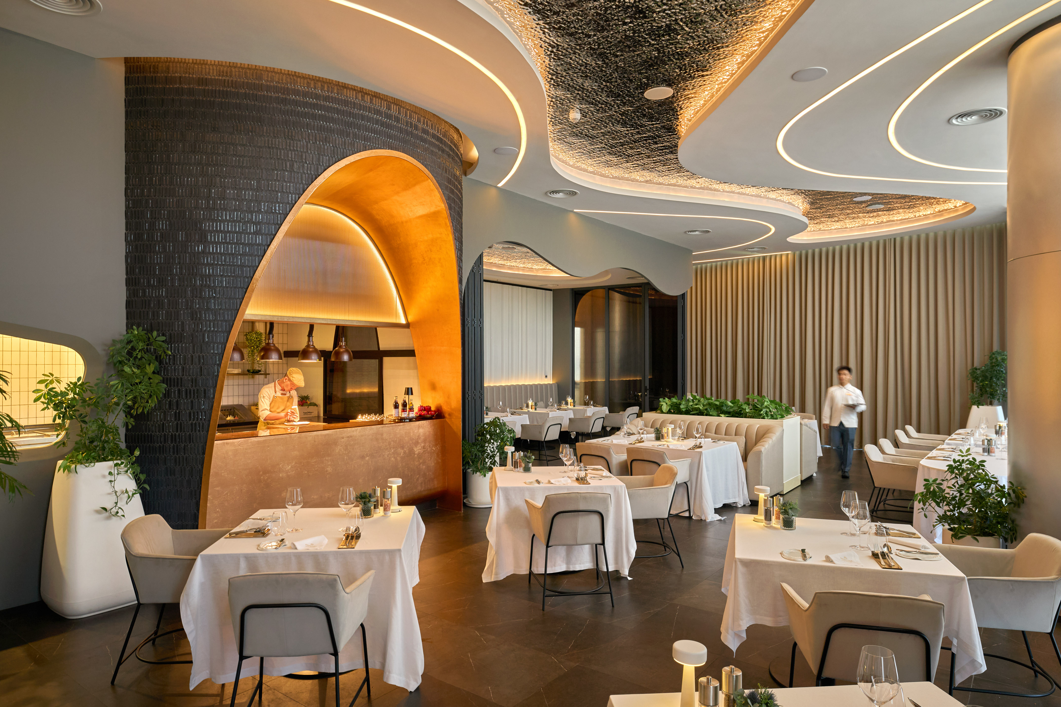 Upscale restaurant dining room with open kitchen, chef, and modern lighting.