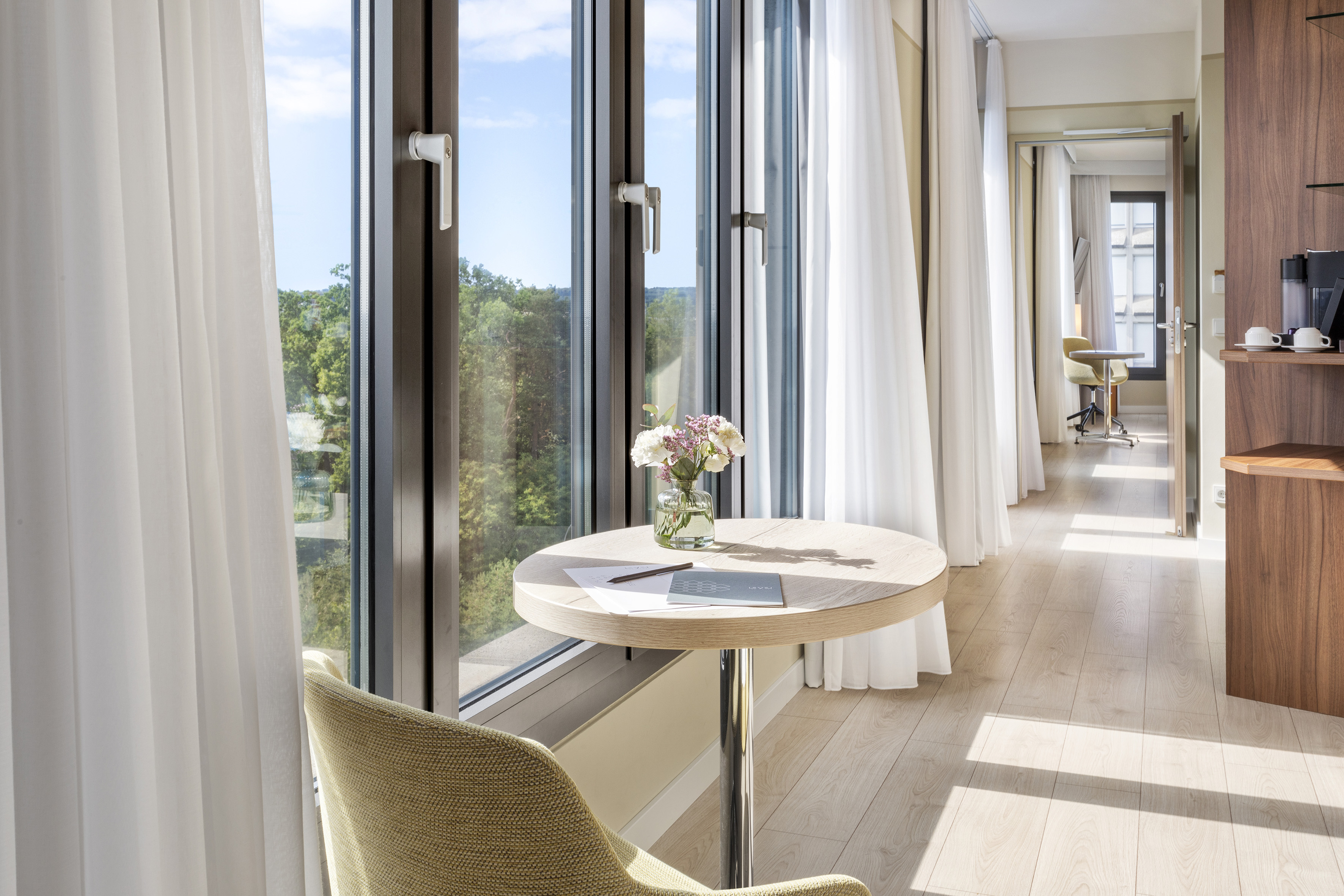 Modern hotel room with large window, city park view, and a small table with flowers.