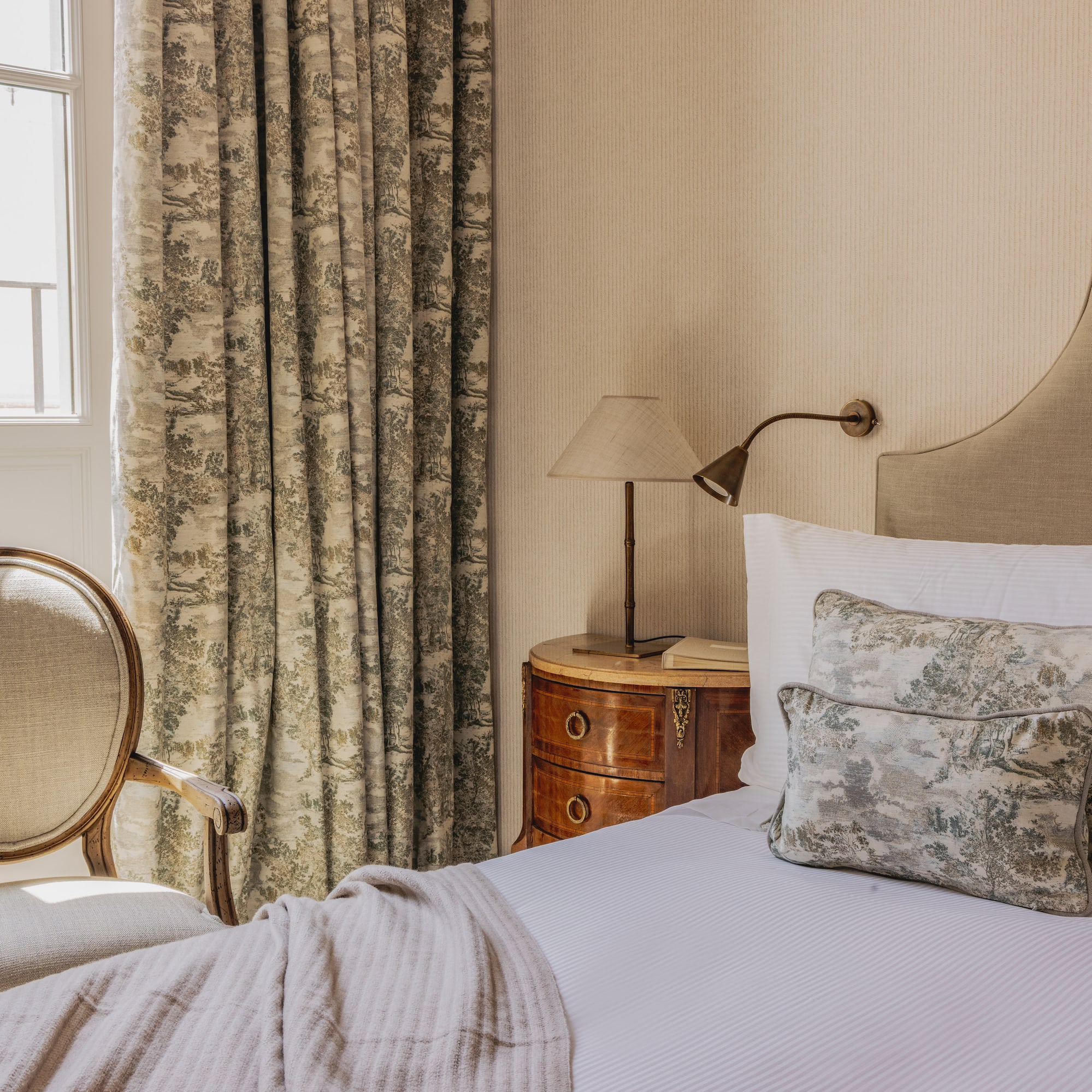 Elegant classic hotel bedroom with upholstered bed, toile curtains, and wooden chair.