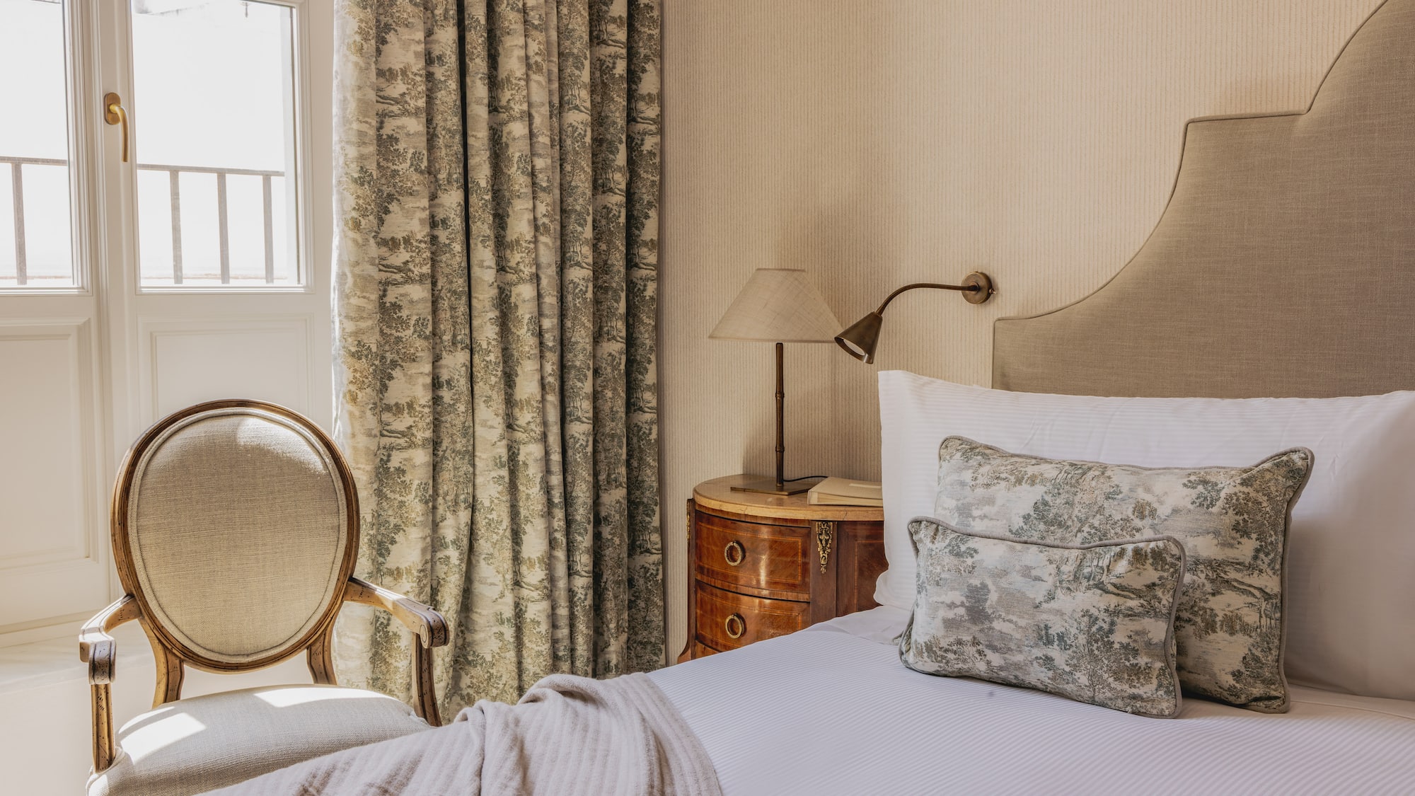 Elegant classic hotel bedroom with upholstered bed, toile curtains, and wooden chair.