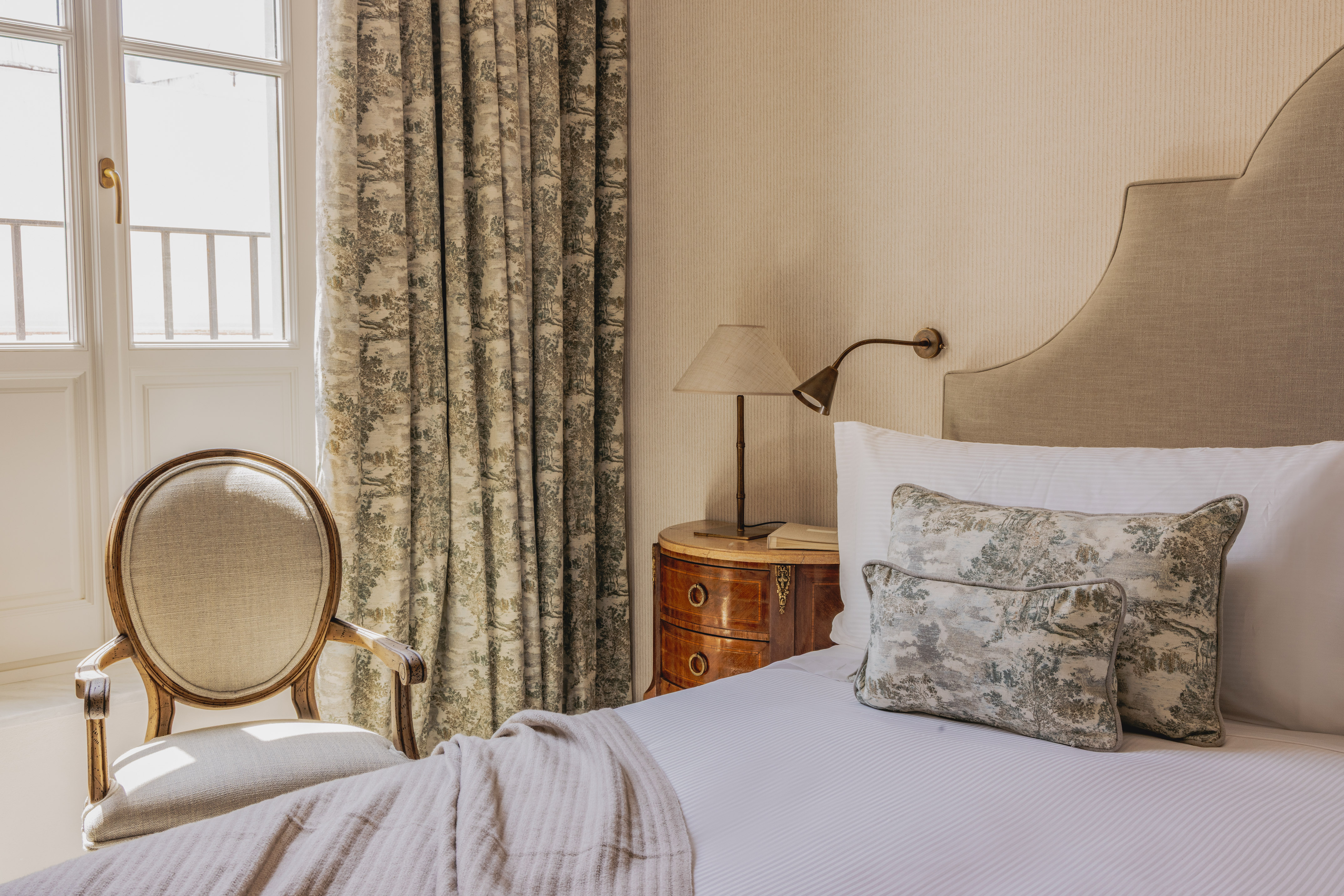 Elegant classic hotel bedroom with upholstered bed, toile curtains, and wooden chair.