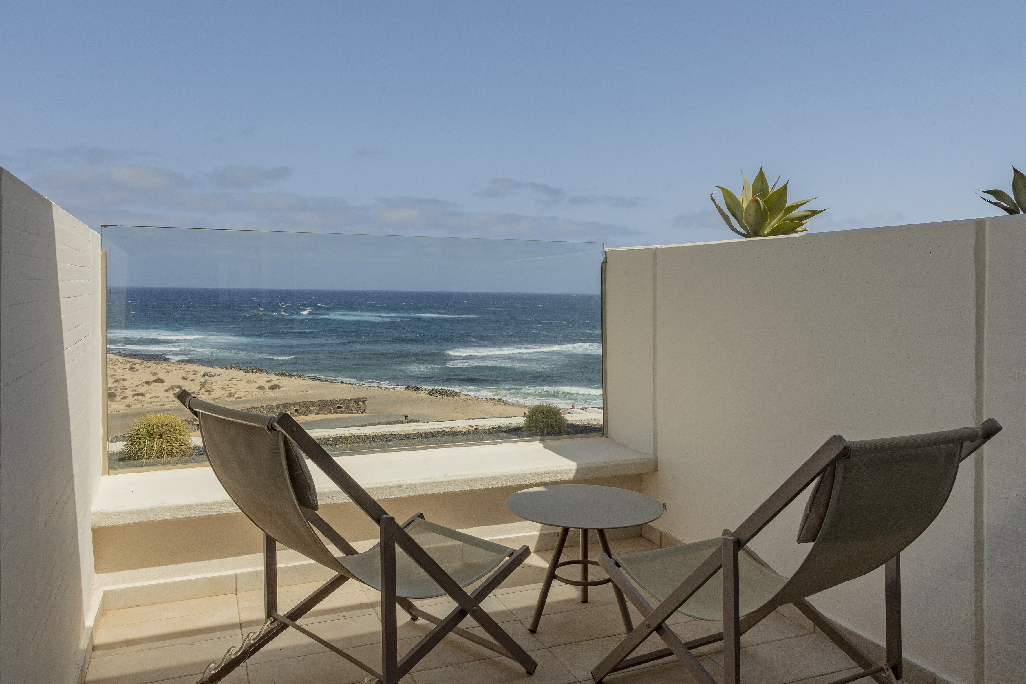 Balcony with ocean view, sandy beach, desert plants, chairs, and table.