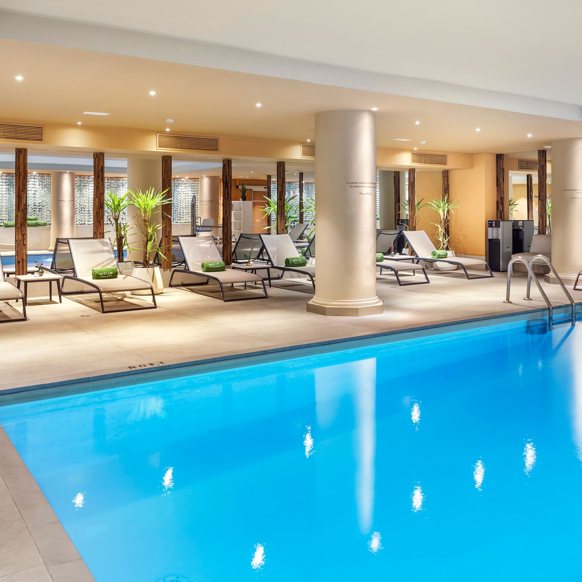 Indoor hotel swimming pool with blue water, lounge chairs, and plants.