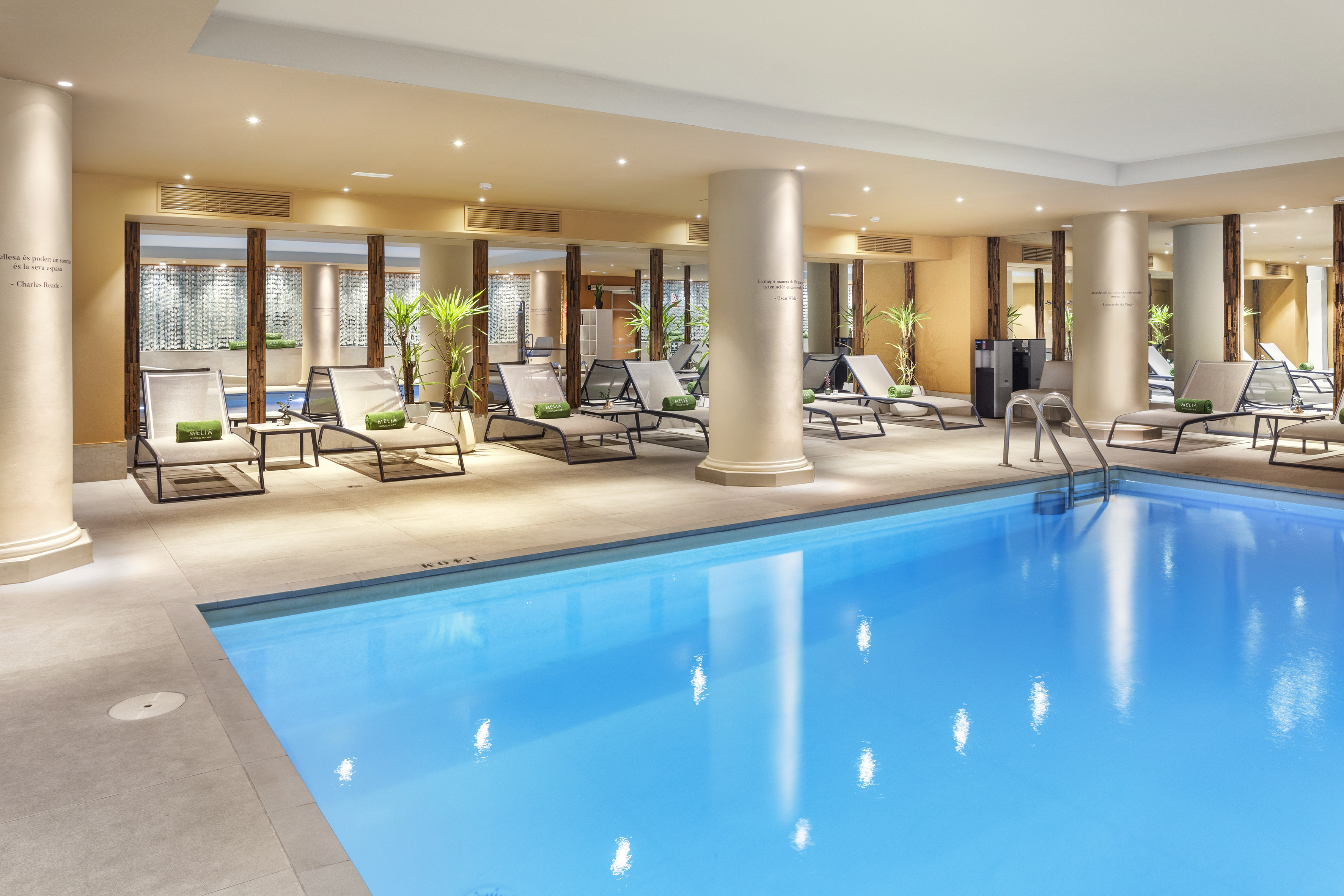 Indoor hotel swimming pool with blue water, lounge chairs, and plants.