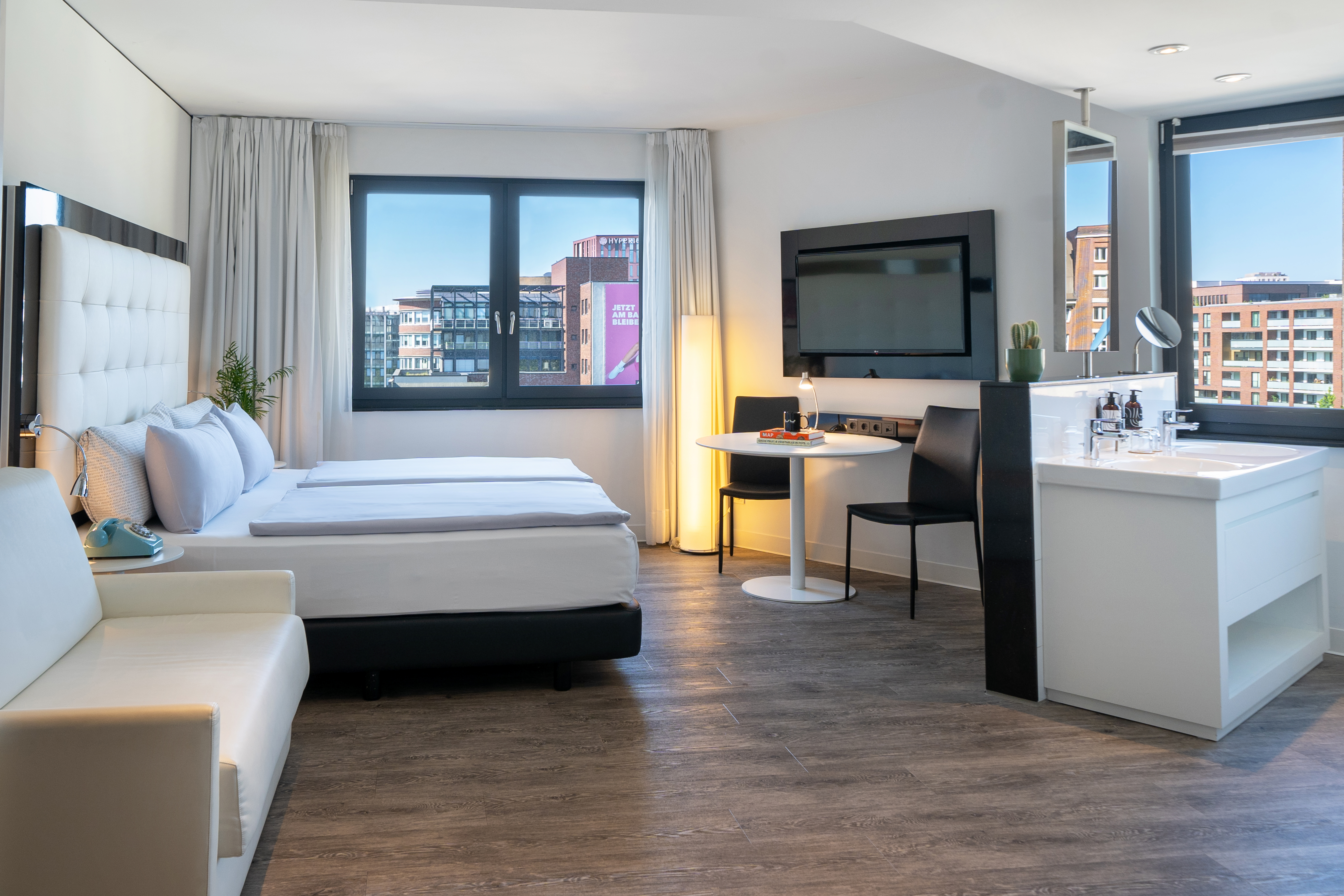 Modern hotel room with bed, city view, wall-mounted TV, and open sink area.