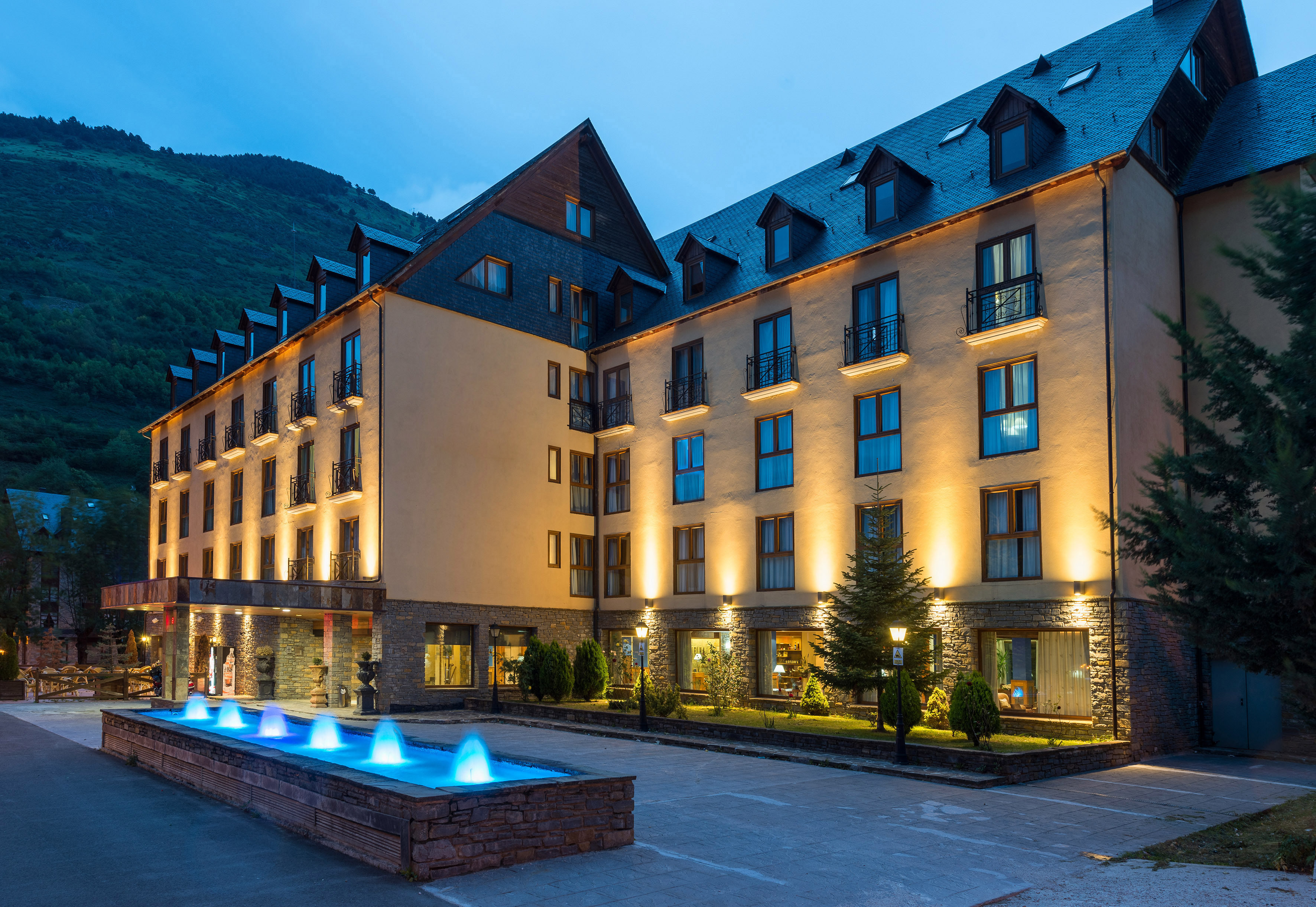 Illuminated mountain hotel with blue fountain at dusk, modern architecture.