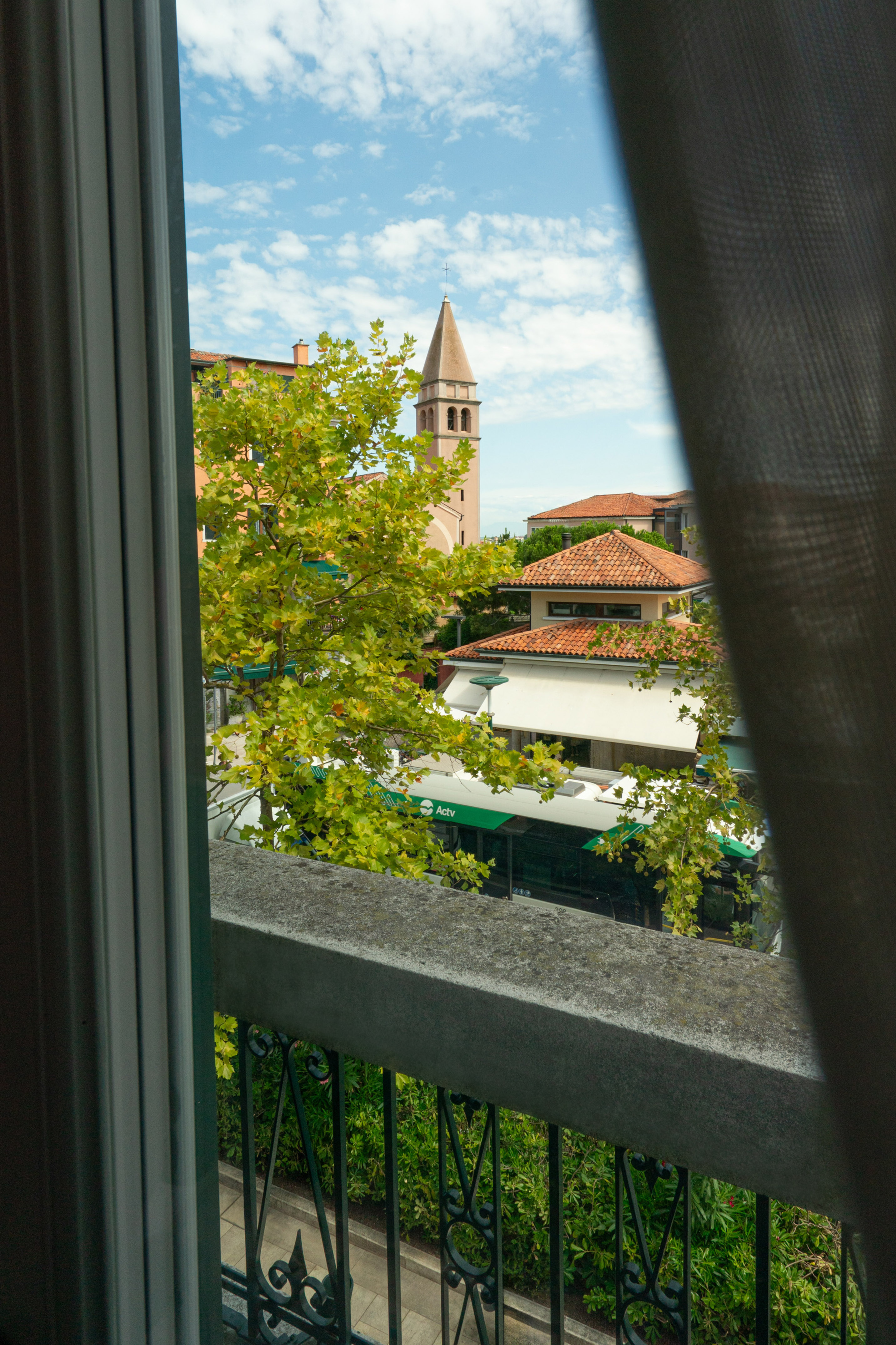 Balcony view: town bell tower, terracotta roofs, green tree, ACTV bus.