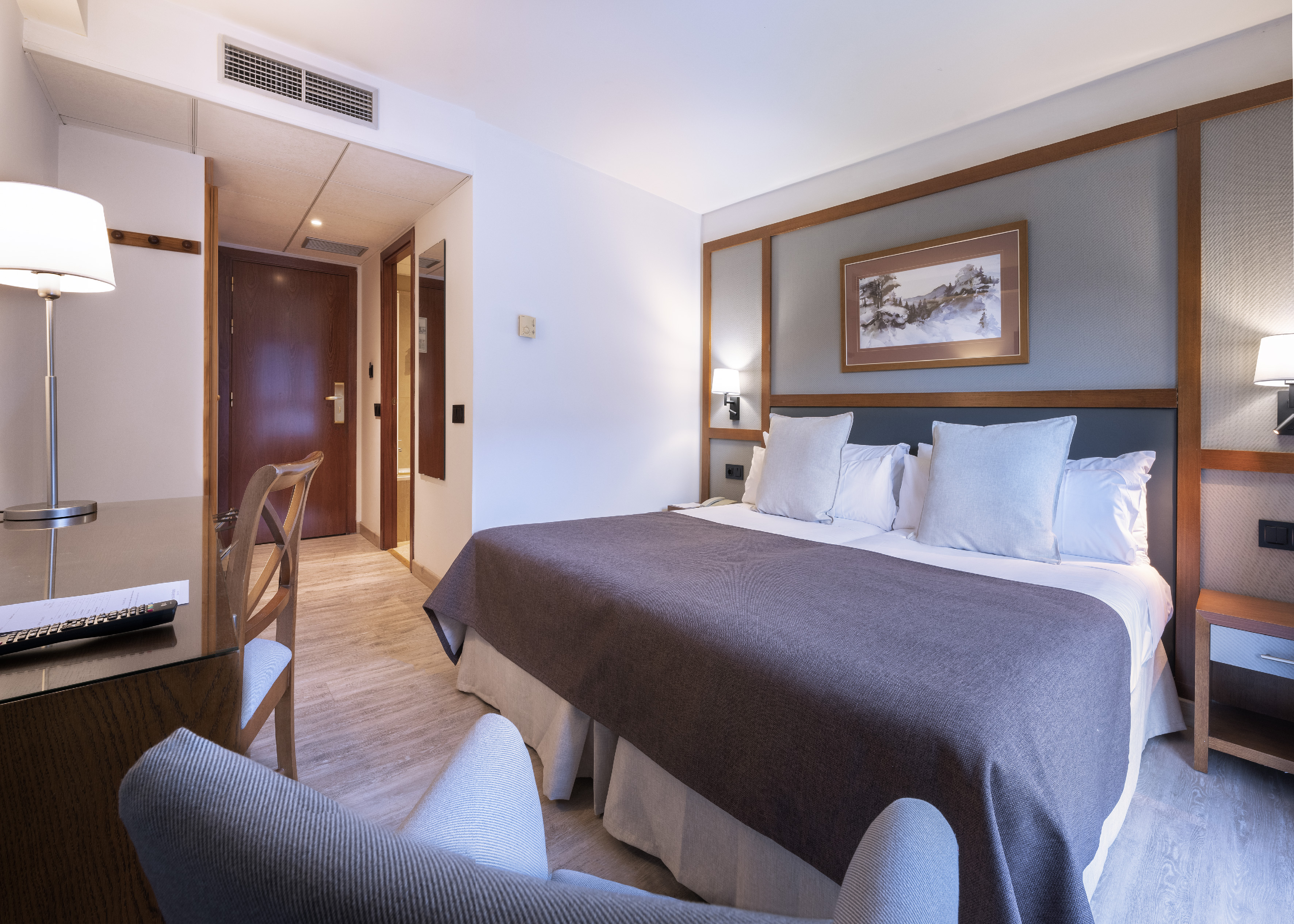 Modern hotel bedroom with large bed, patterned headboard, desk, and armchair.