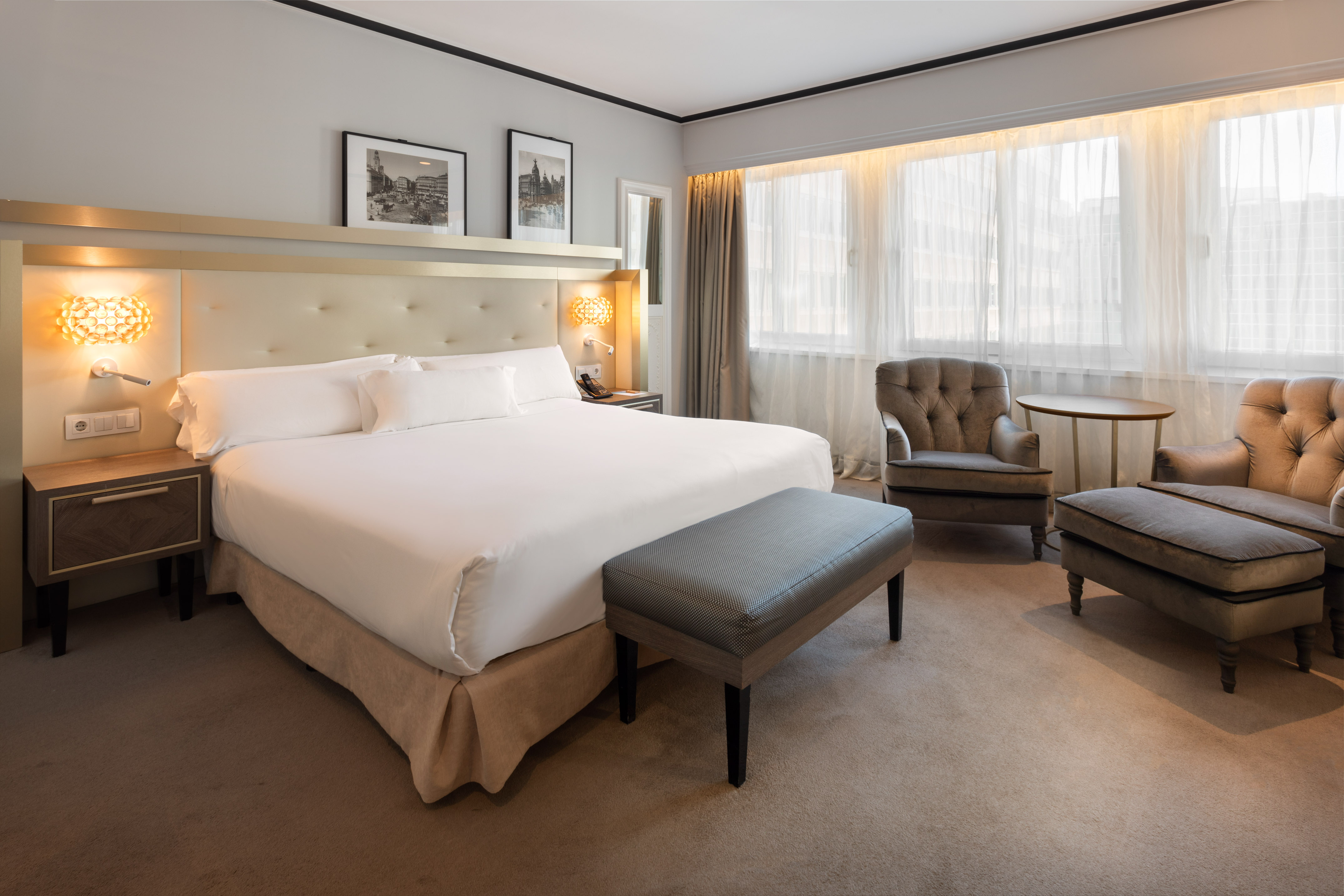 Luxury hotel room with king bed, beige headboard, armchairs, and bright windows.