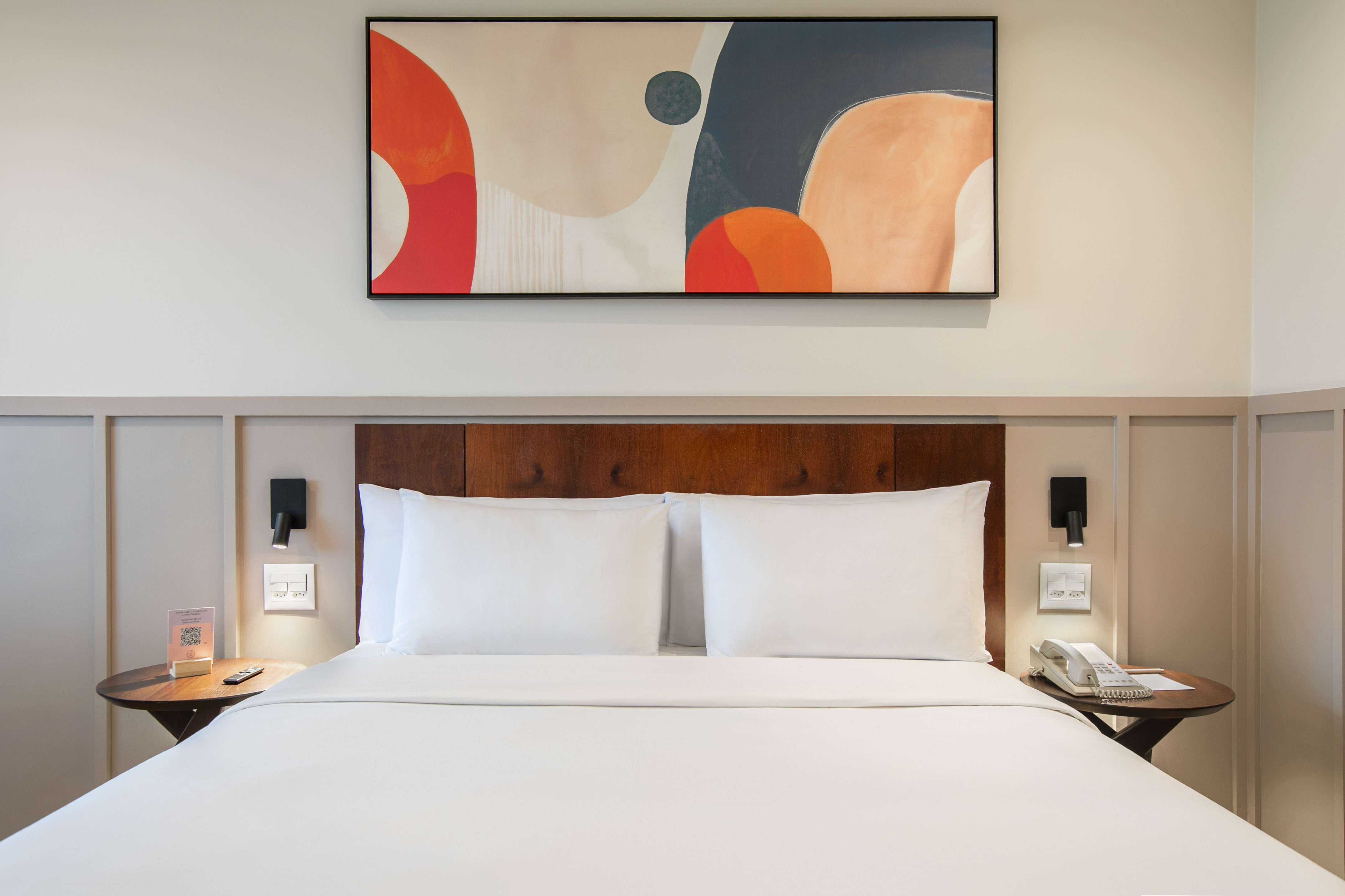 Modern hotel room bed, white linens, wooden headboard, abstract art.