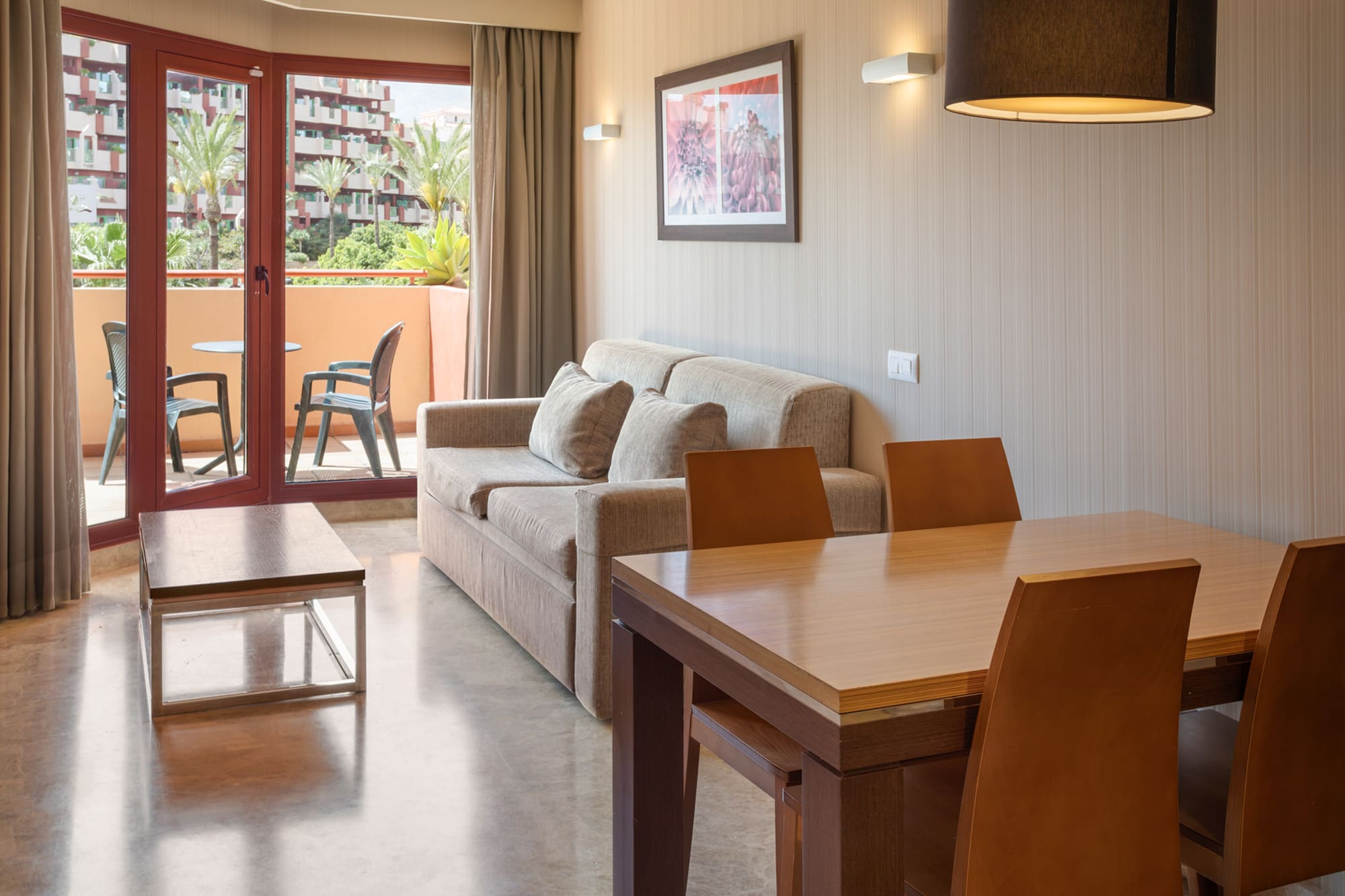 Hotel apartment living room with sofa, dining table, and balcony overlooking palm trees.