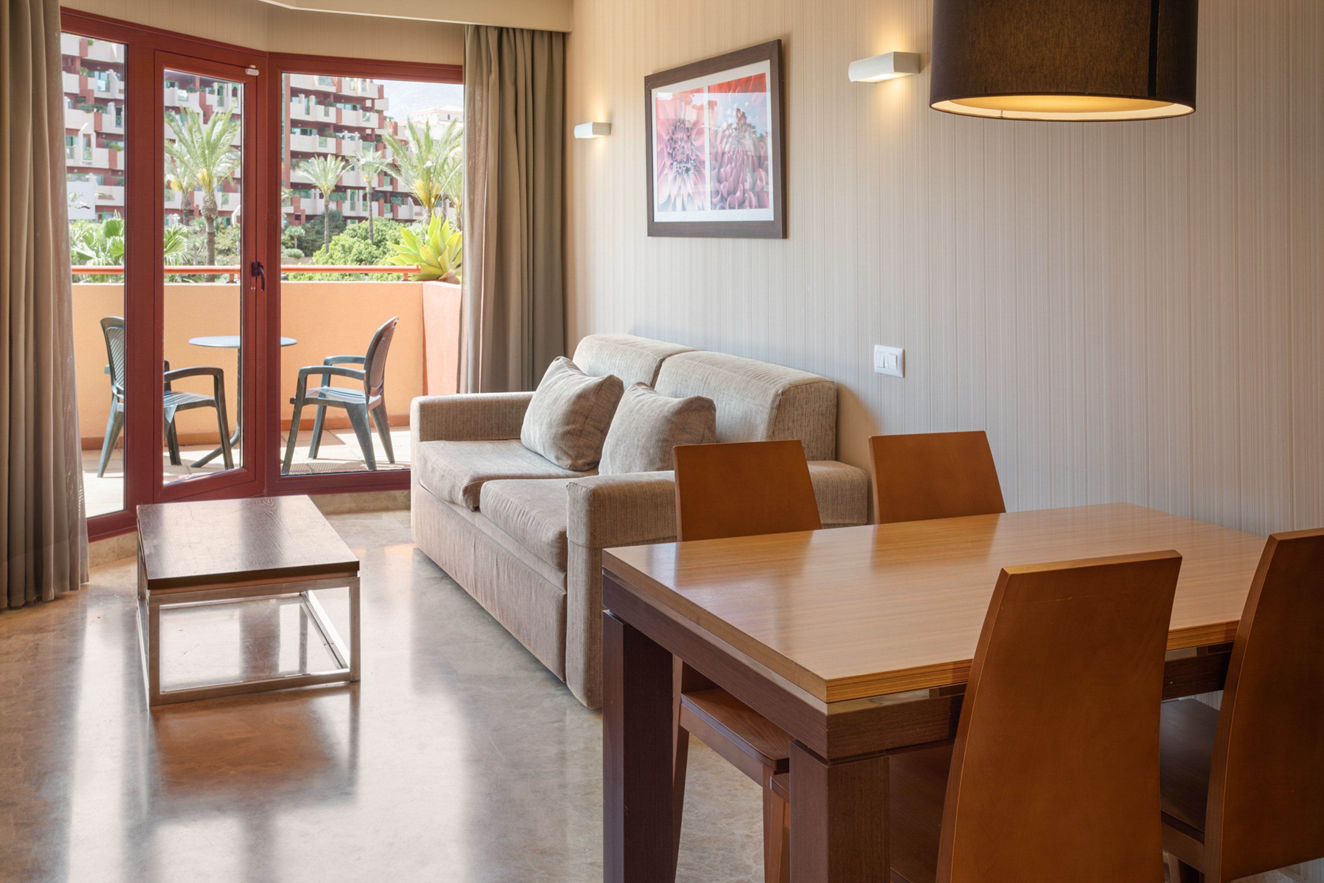 Hotel apartment living room with sofa, dining table, and balcony overlooking palm trees.