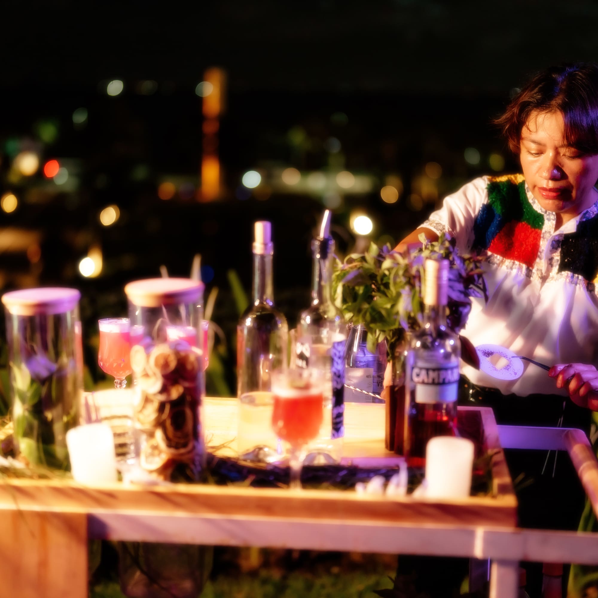 a woman standing at a table with bottles and a drink