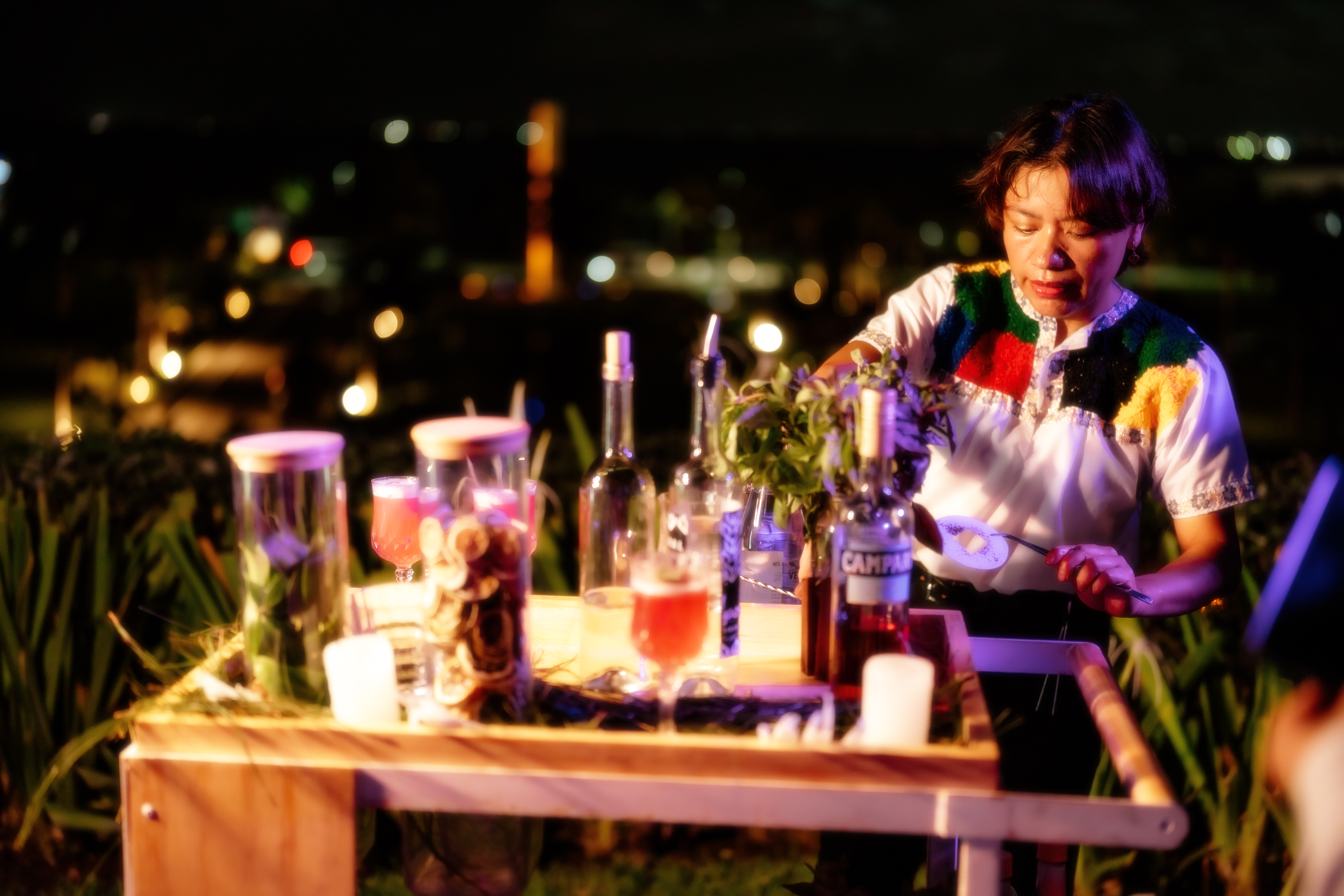 a woman standing at a table with bottles and a drink