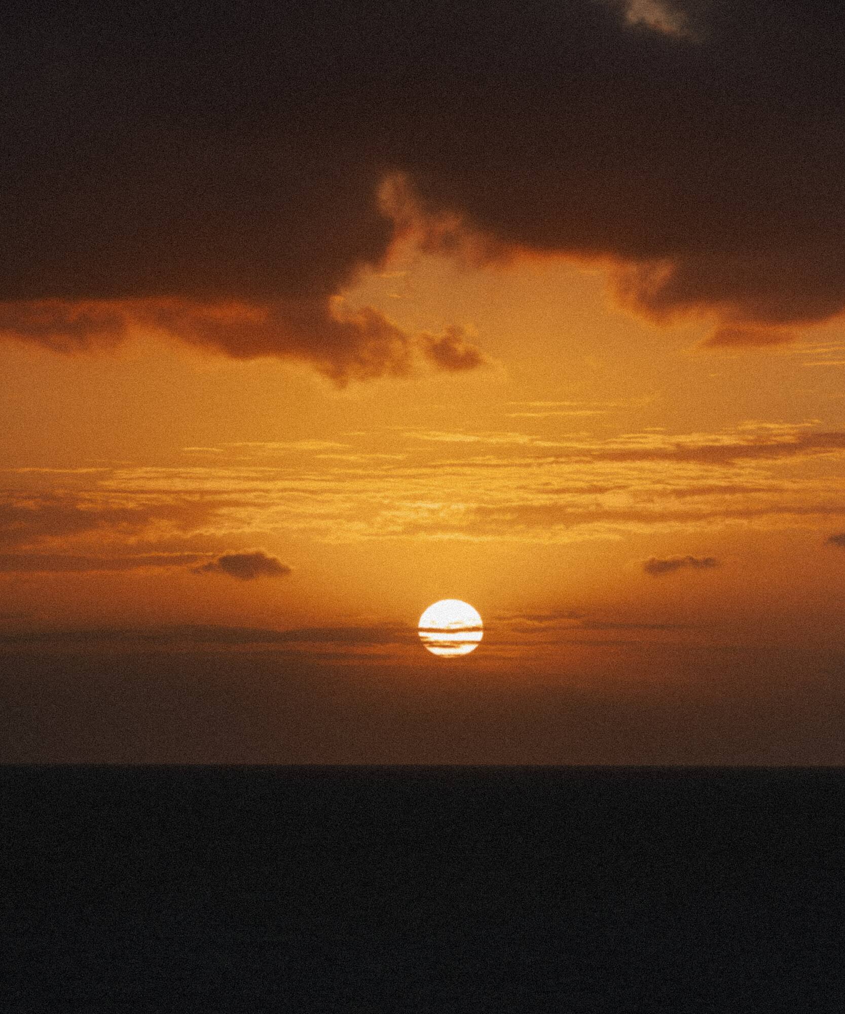 Atardecer naranja sobre el mar, sol poniente y nubes oscuras en el cielo.