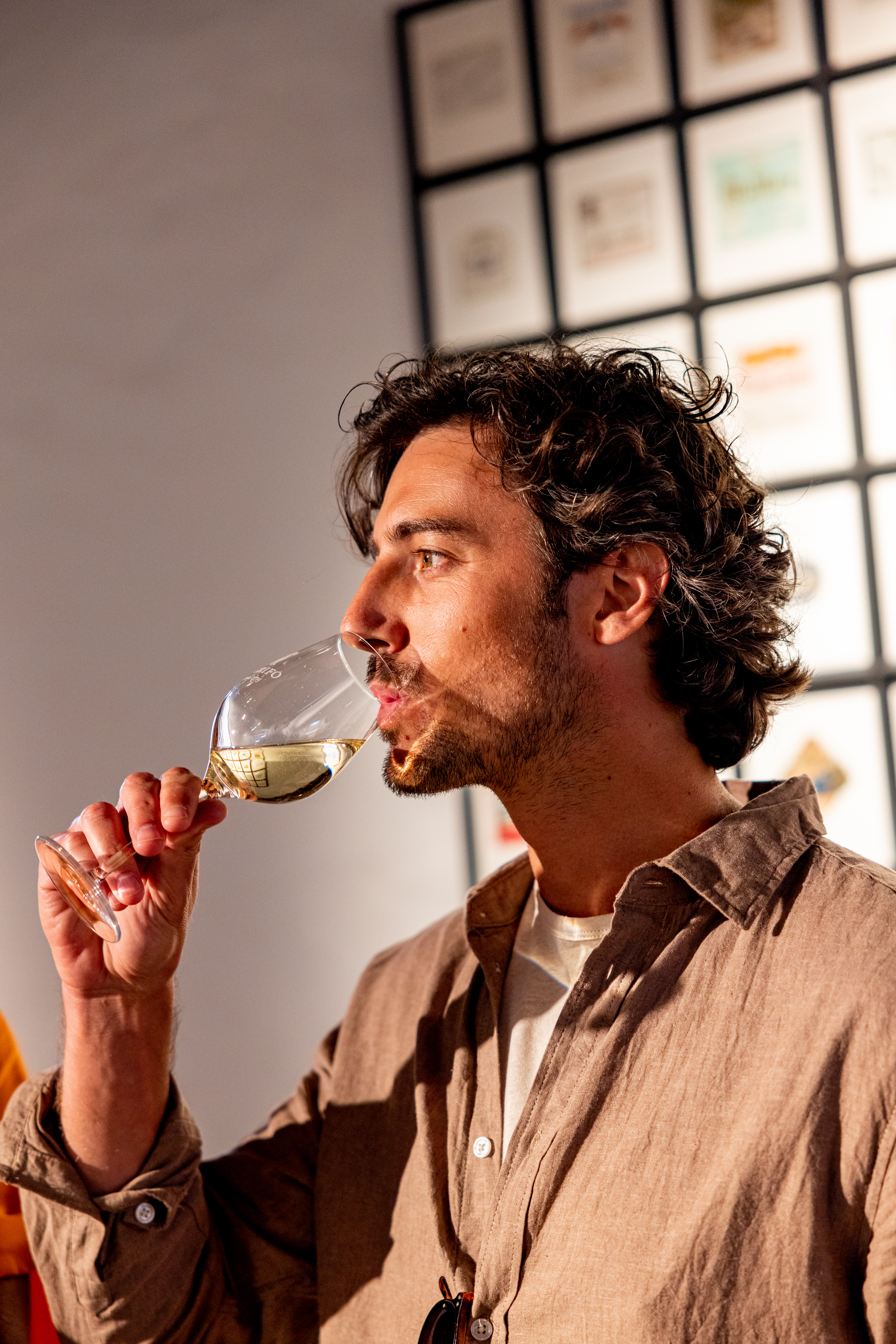 Man with curly hair tasting white wine from a glass at an event.