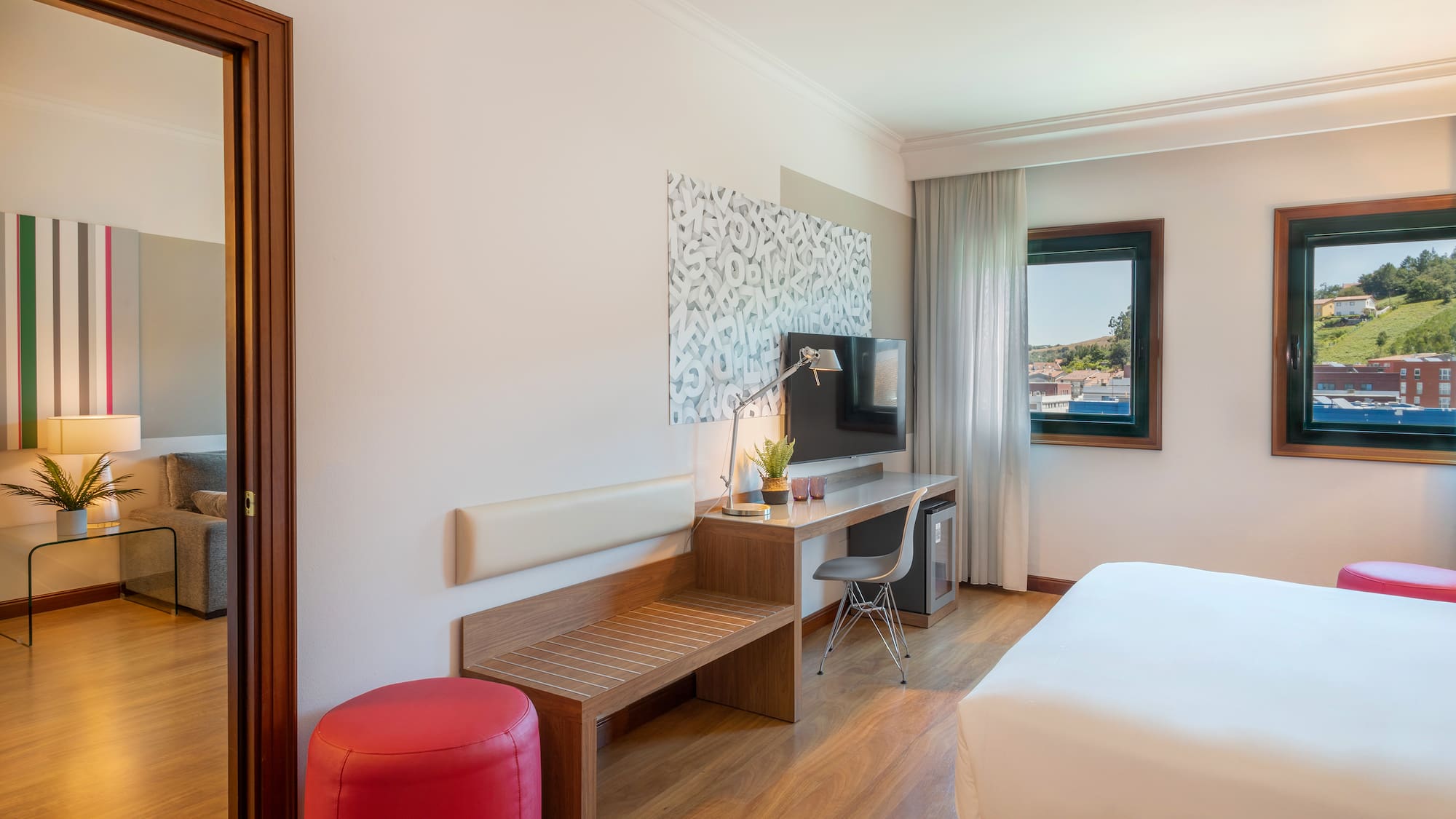 Modern hotel room with bed, desk, TV, wooden floor, and scenic window view.