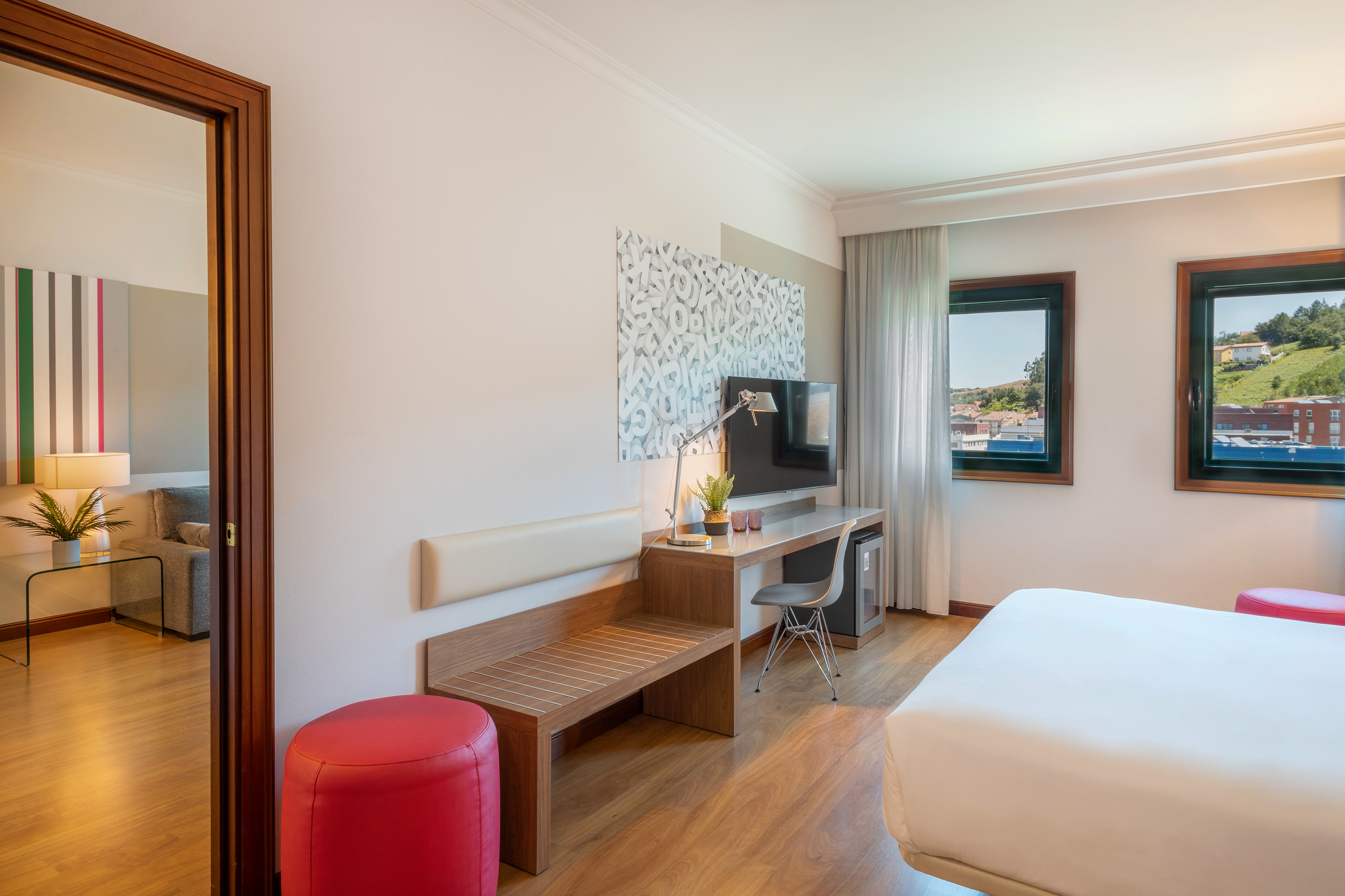 Modern hotel room with bed, desk, TV, wooden floor, and scenic window view.