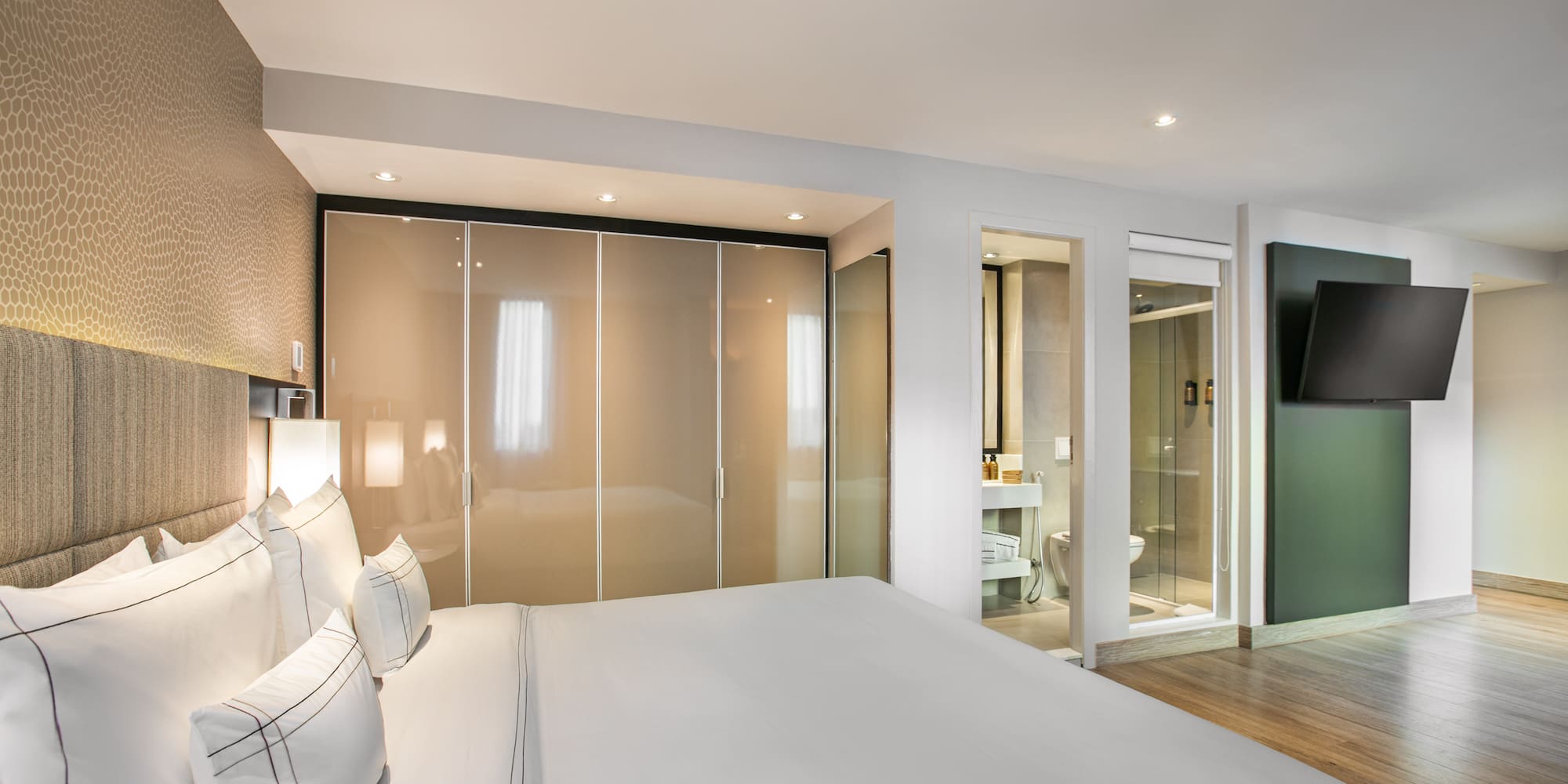 Modern hotel bedroom with white bed, closet, en-suite bath, and wall TV.