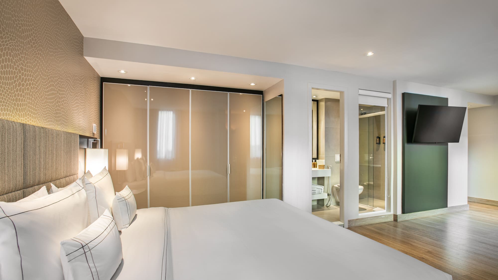 Modern hotel bedroom with white bed, closet, en-suite bath, and wall TV.