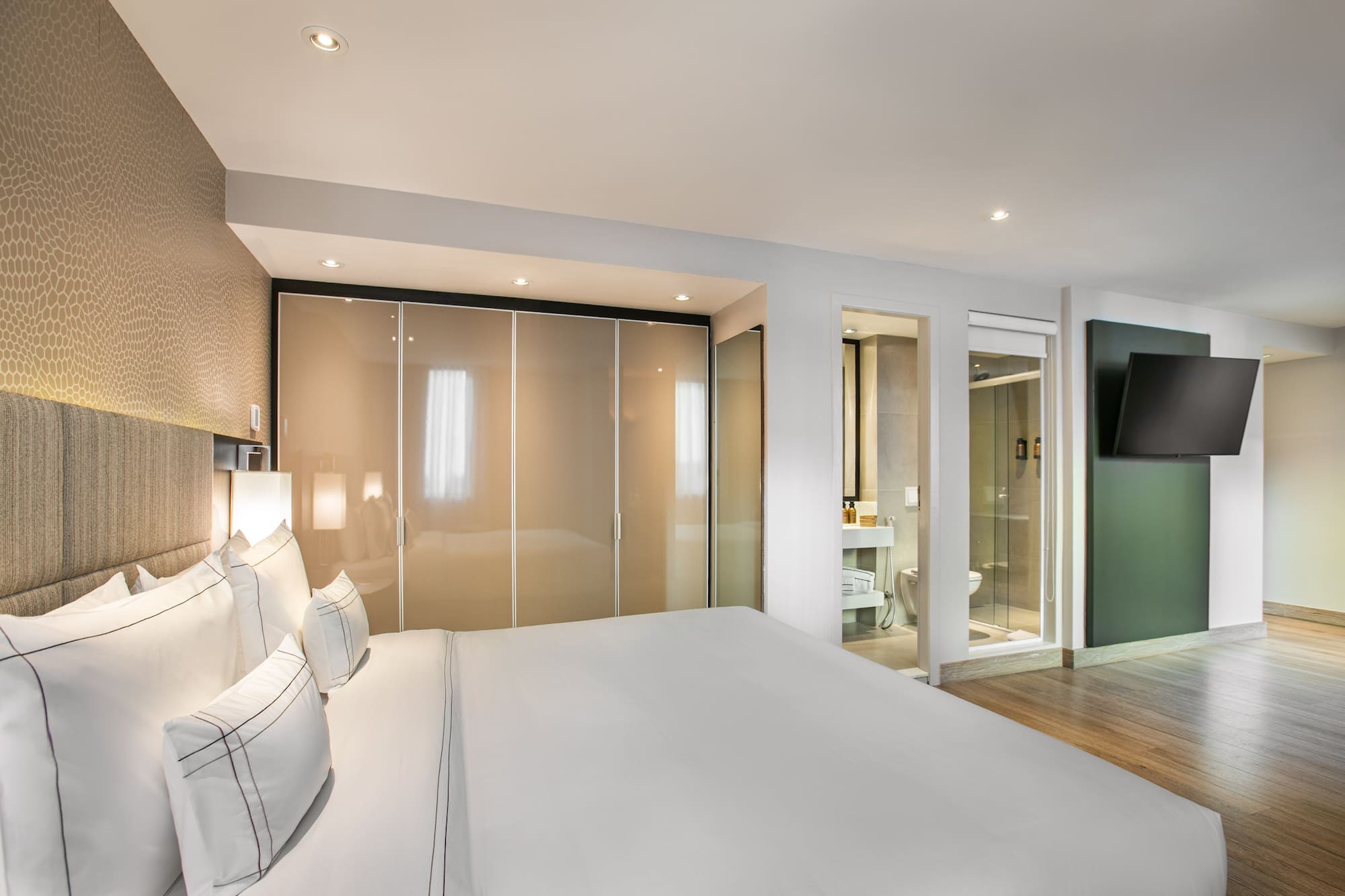 Modern hotel bedroom with white bed, closet, en-suite bath, and wall TV.