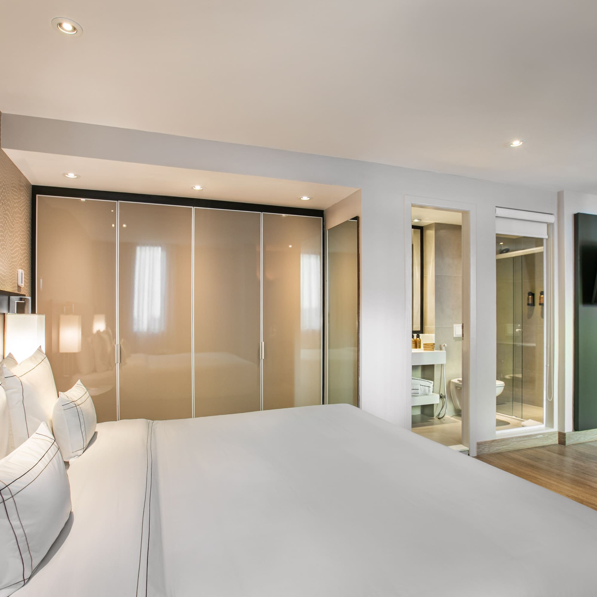 Modern hotel bedroom with white bed, closet, en-suite bath, and wall TV.