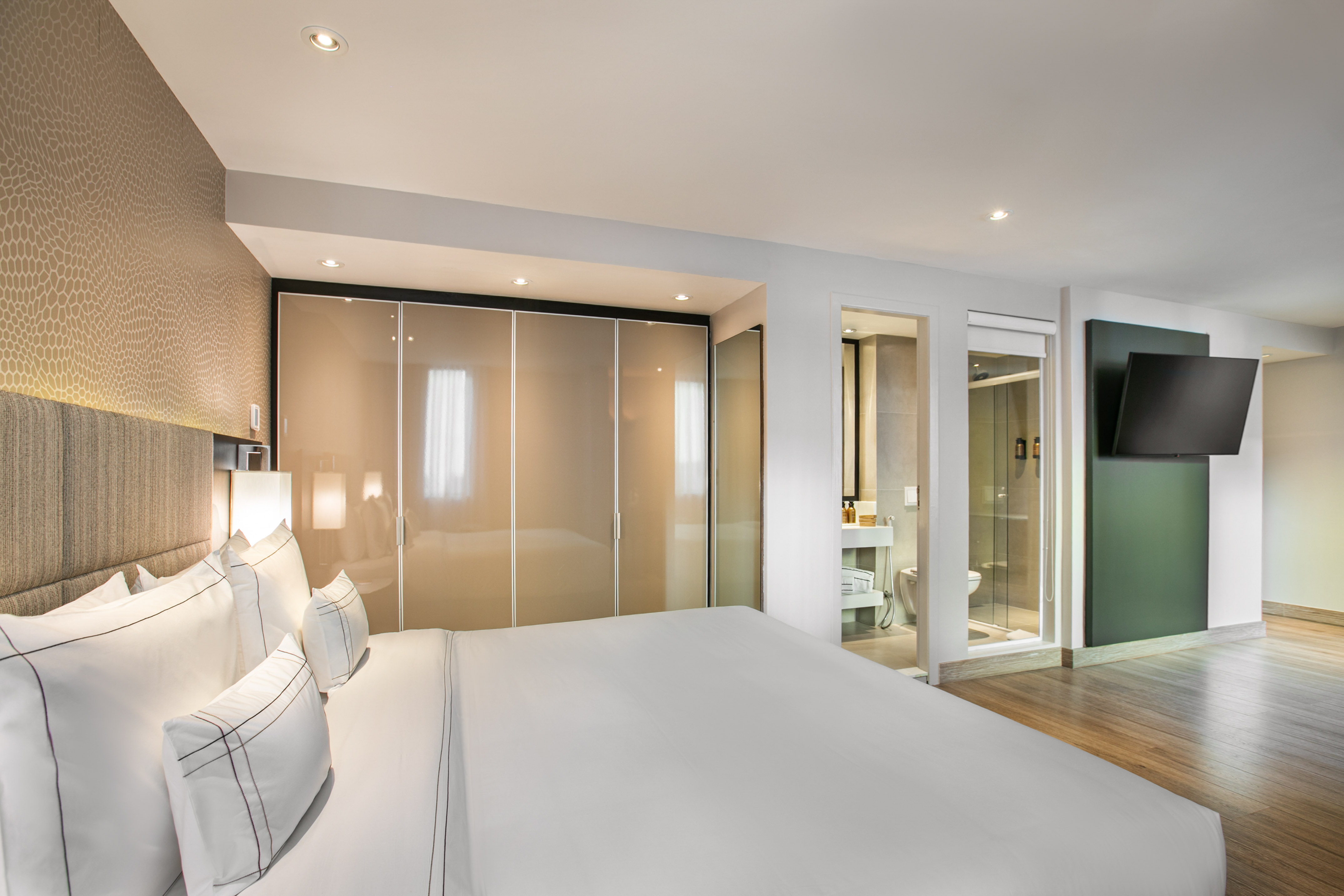Modern hotel bedroom with white bed, closet, en-suite bath, and wall TV.