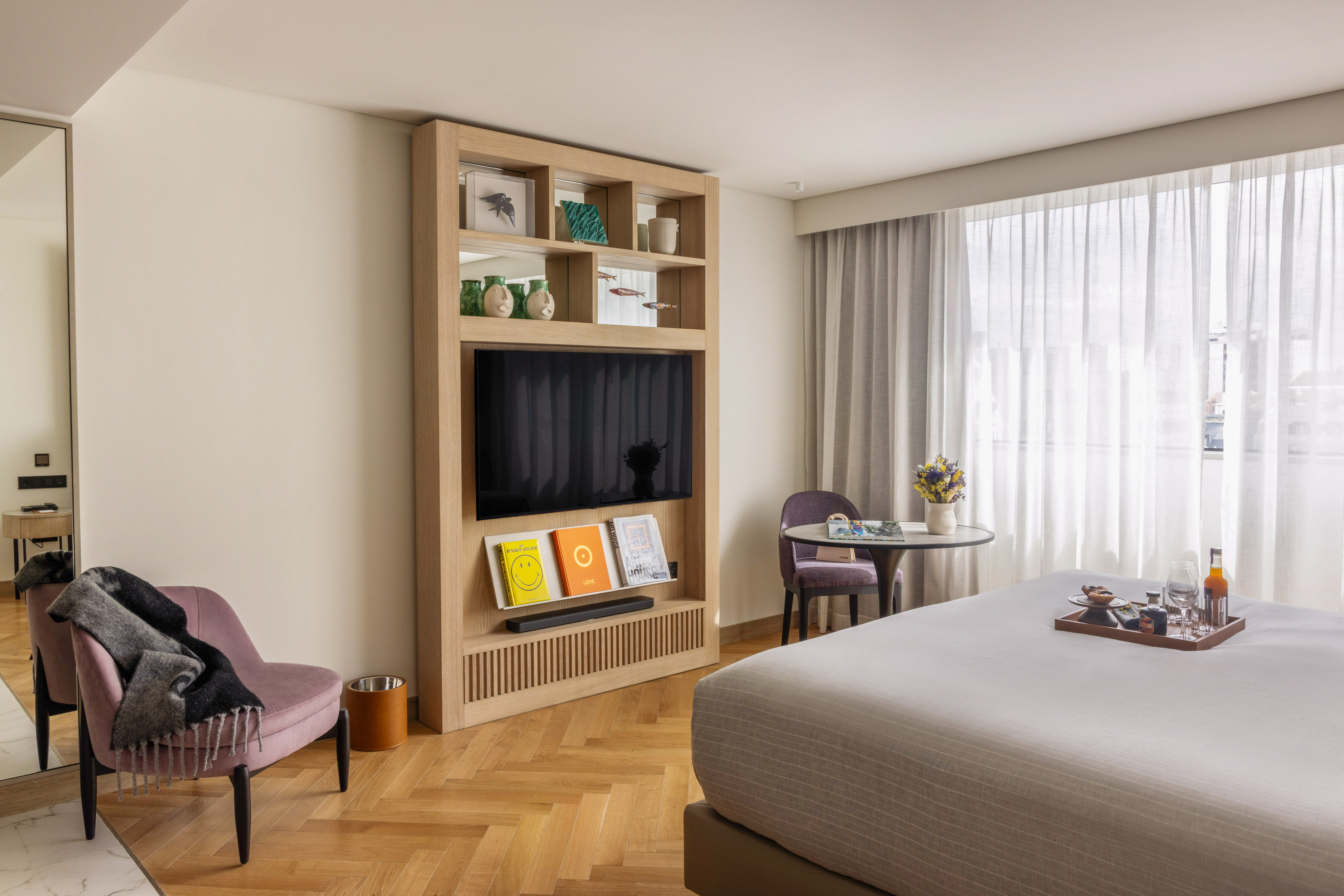 Modern hotel bedroom with TV, large bed, armchair, and hardwood floor.