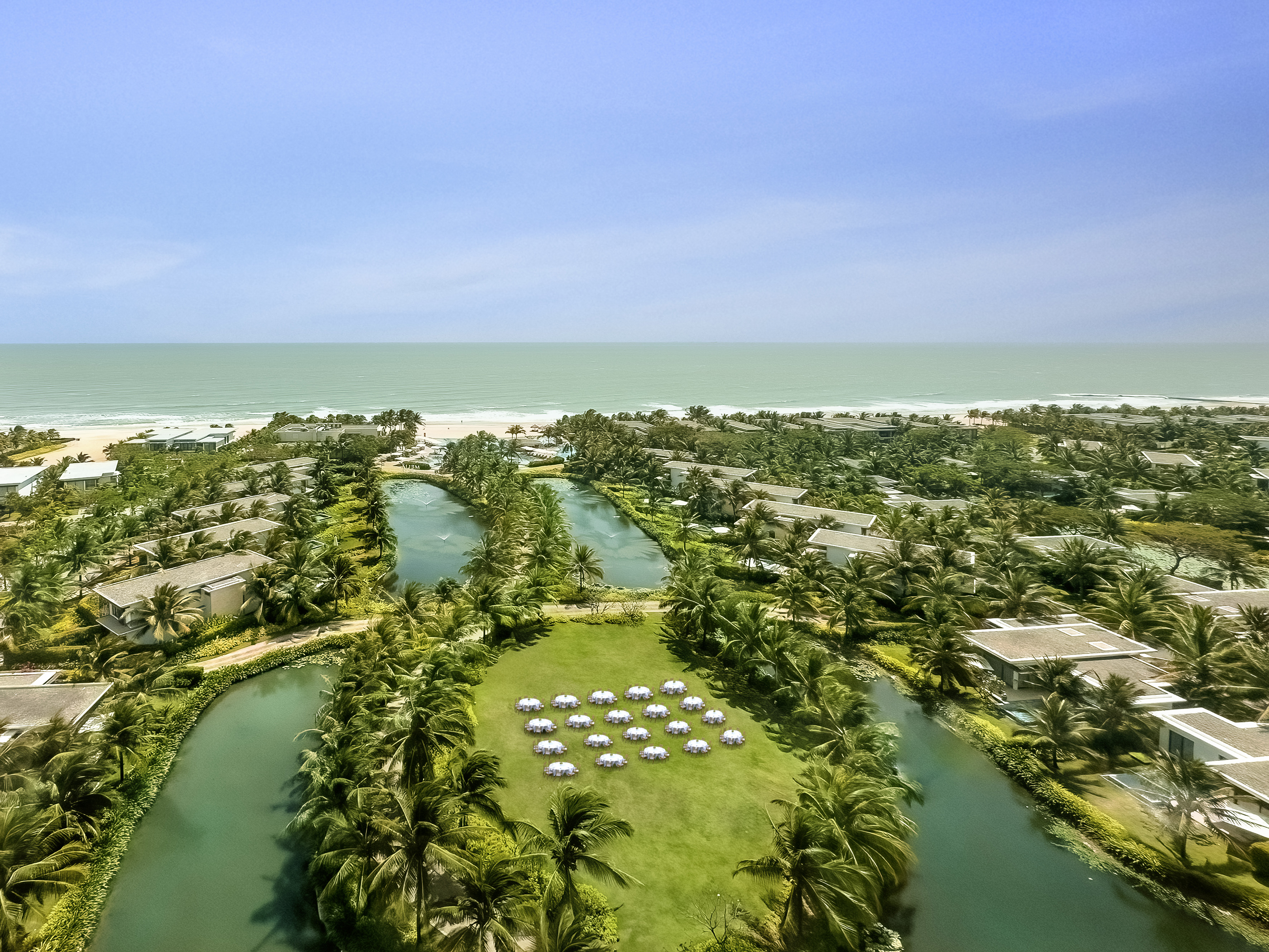 Aerial tropical resort with beach, ocean, palm trees, canals, and event setup.
