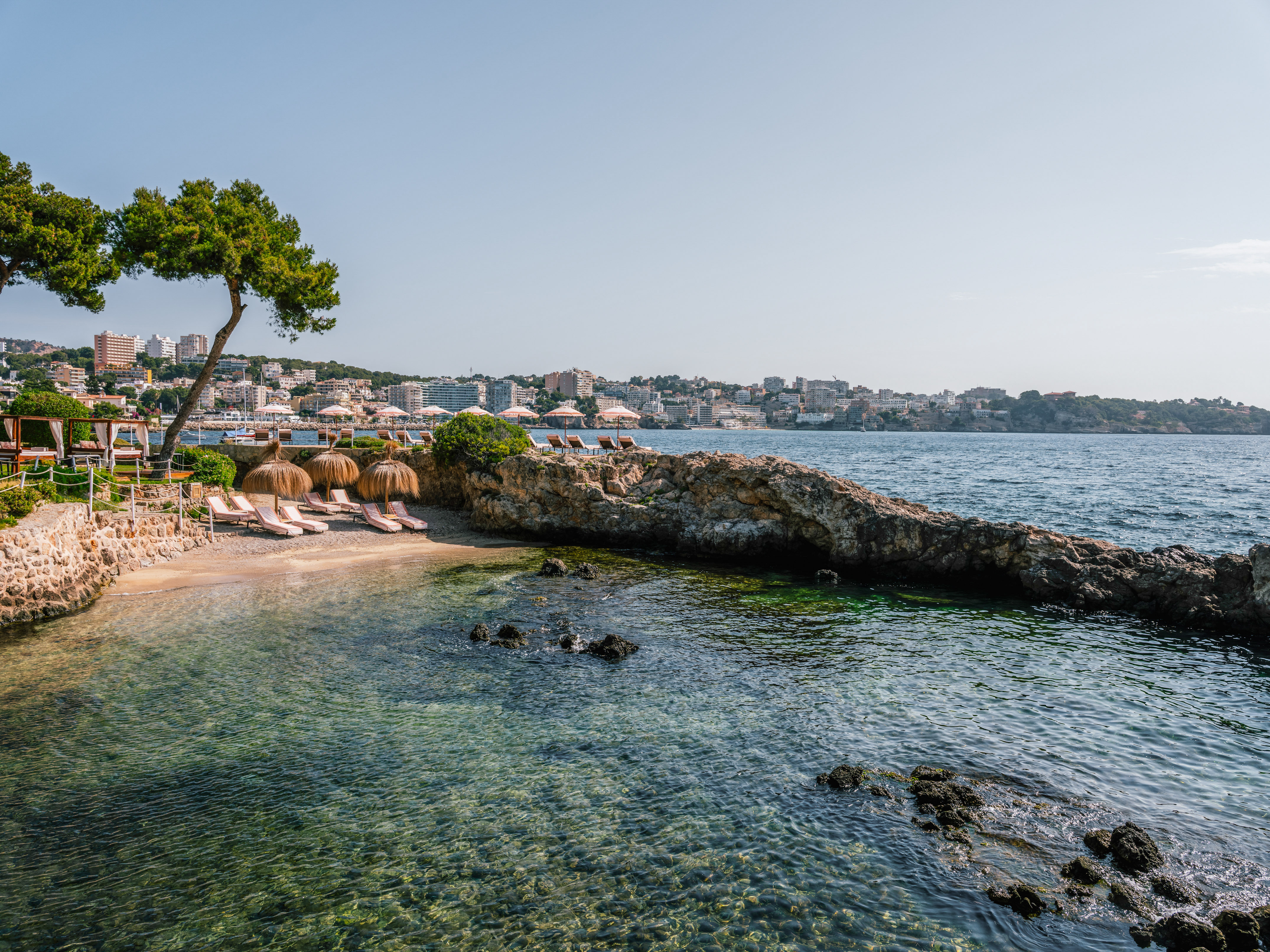 Sunny beach cove with clear water, straw umbrellas, sunbeds, and coastal town.