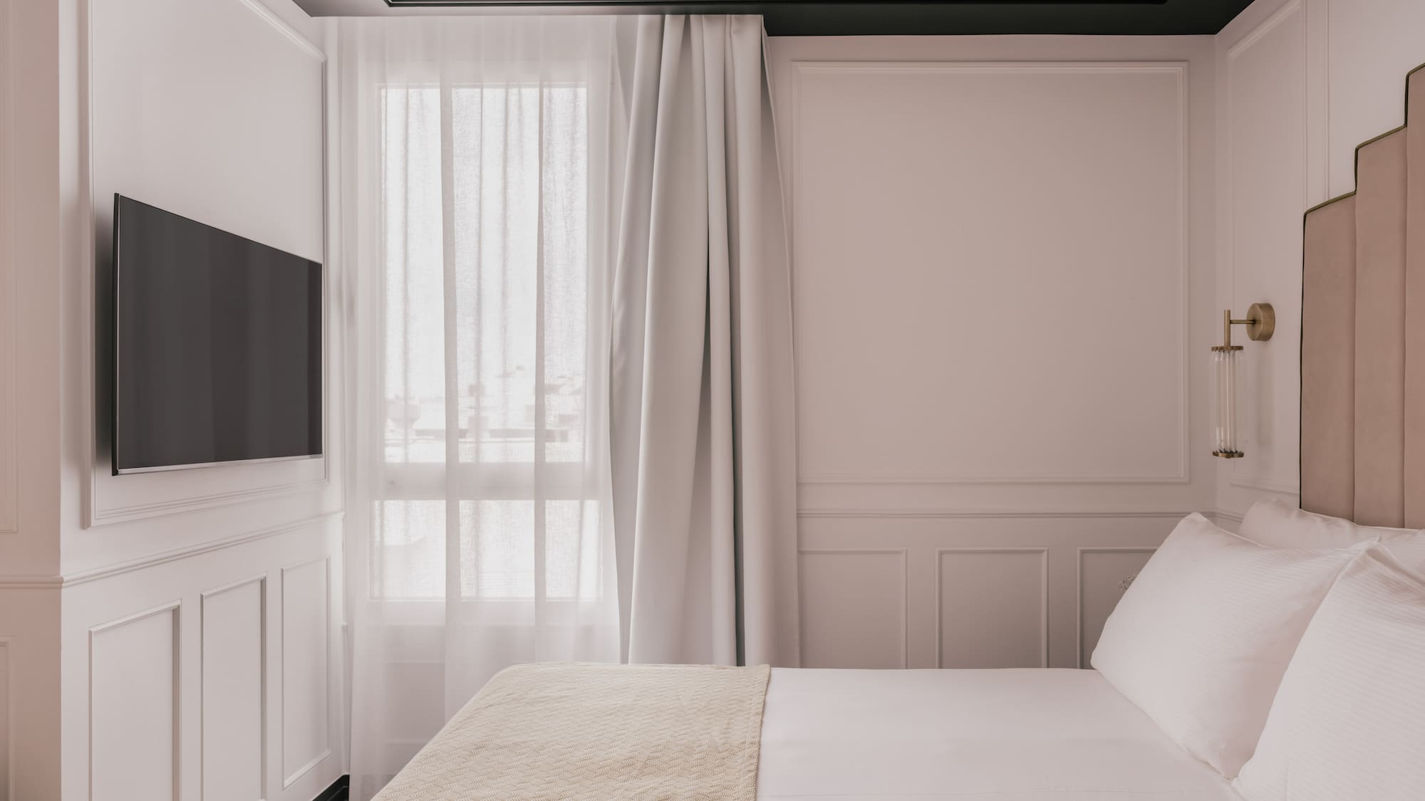 Contemporary bedroom with white paneled walls, beige bed, wall TV, and window.