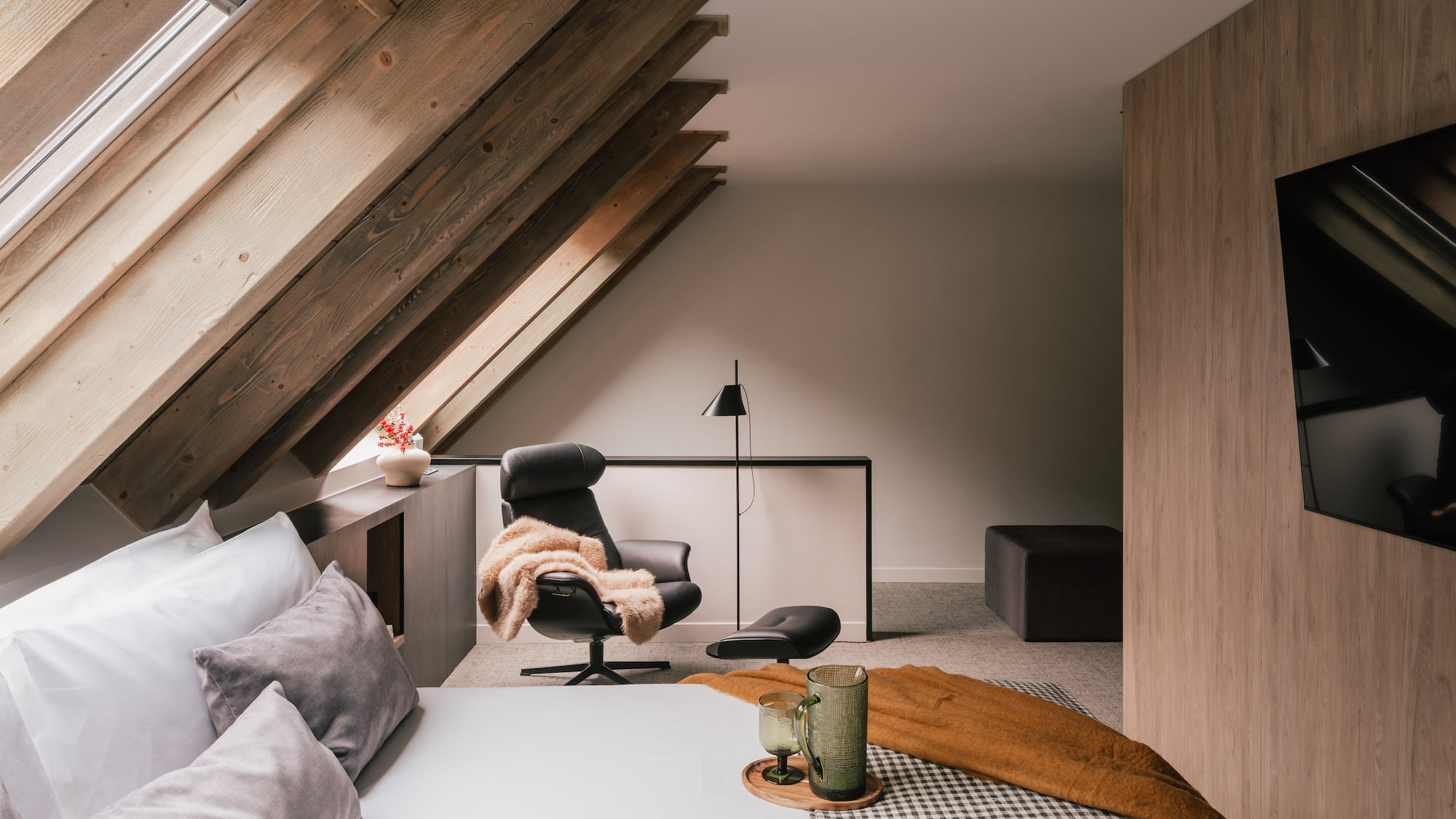Cozy attic bedroom with exposed wooden beams, skylight, bed, and armchair.