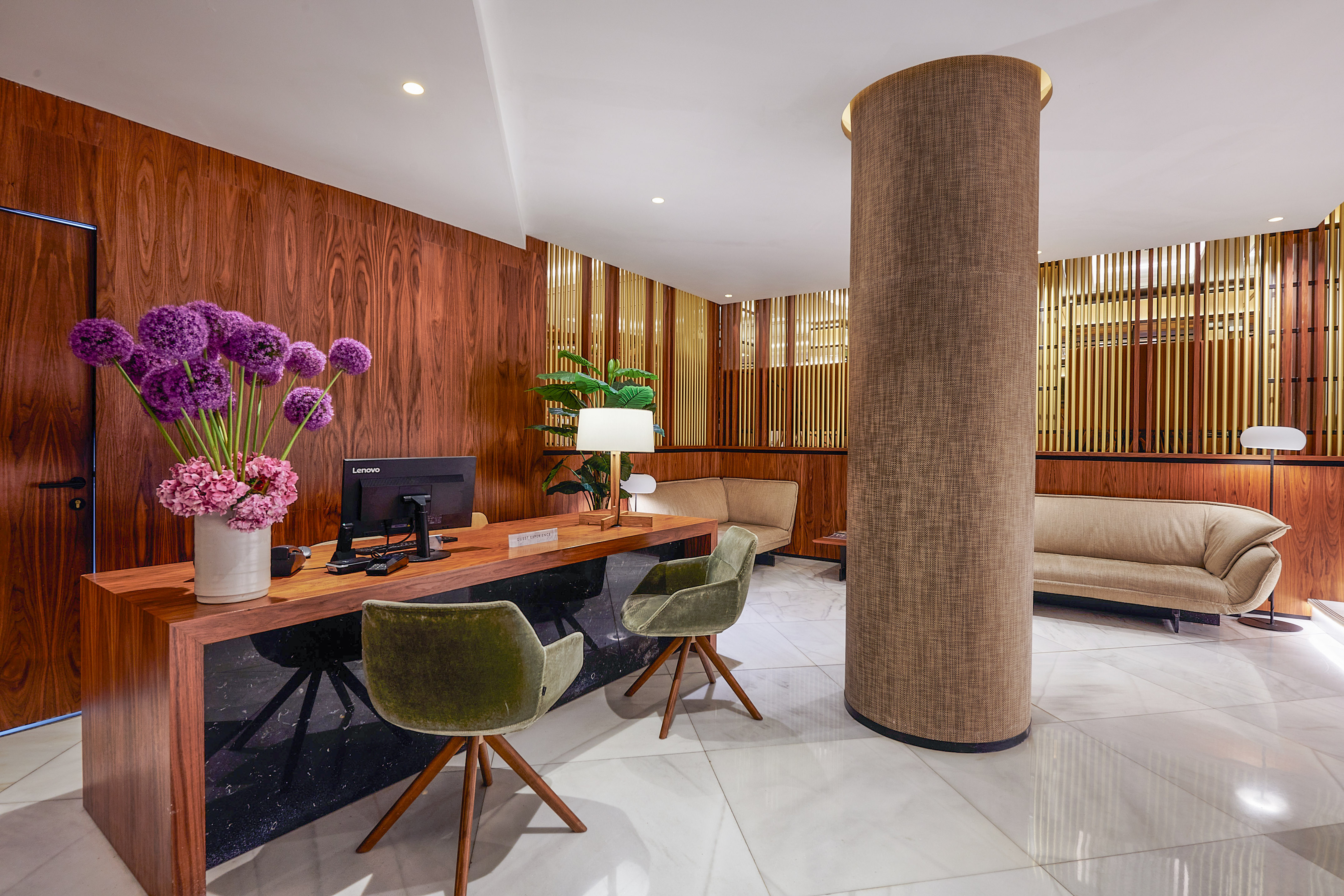 Hotel reception area with wood walls, marble floor, desk, and green chairs.