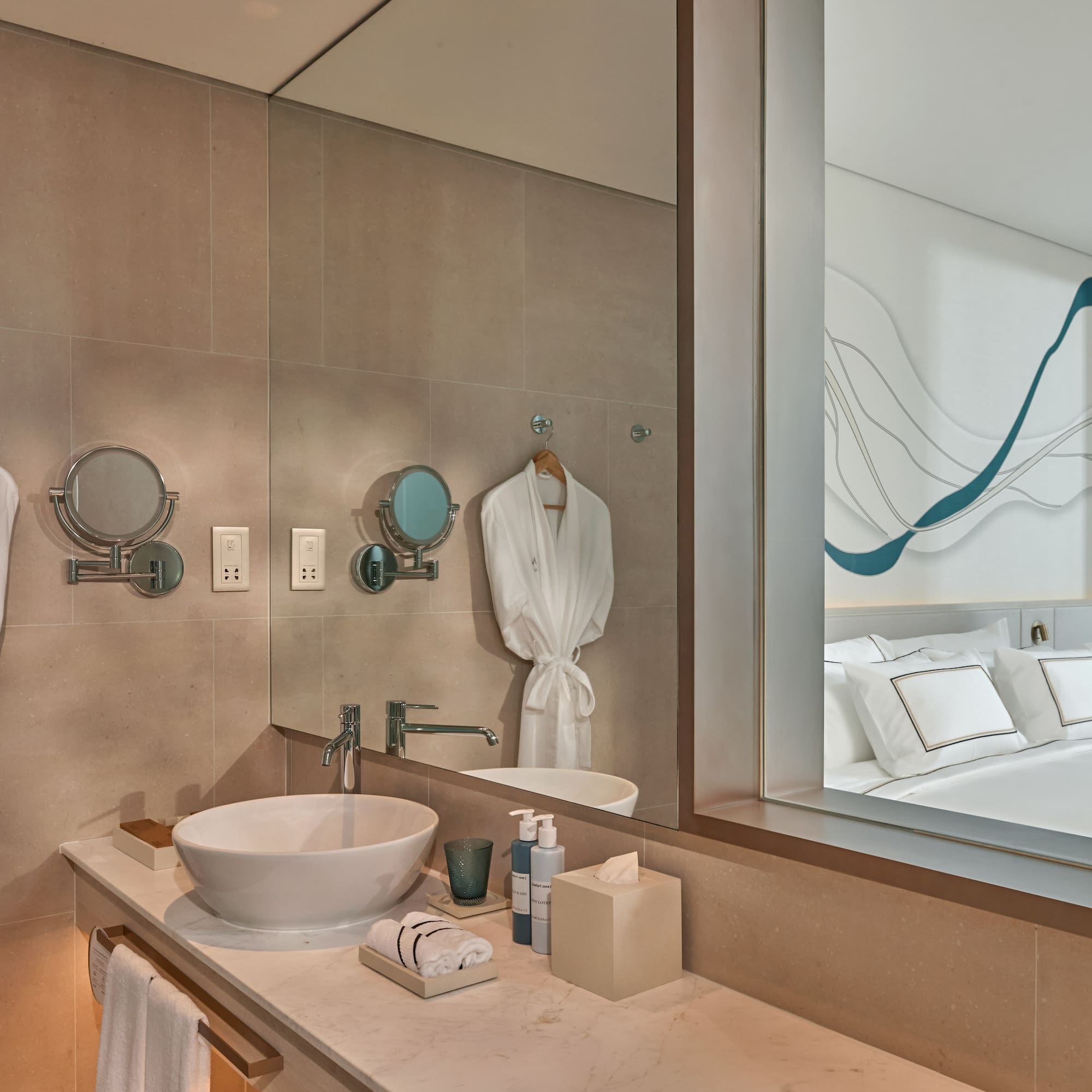 Hotel bathroom vanity with white sink, bathrobes, reflecting modern bedroom.