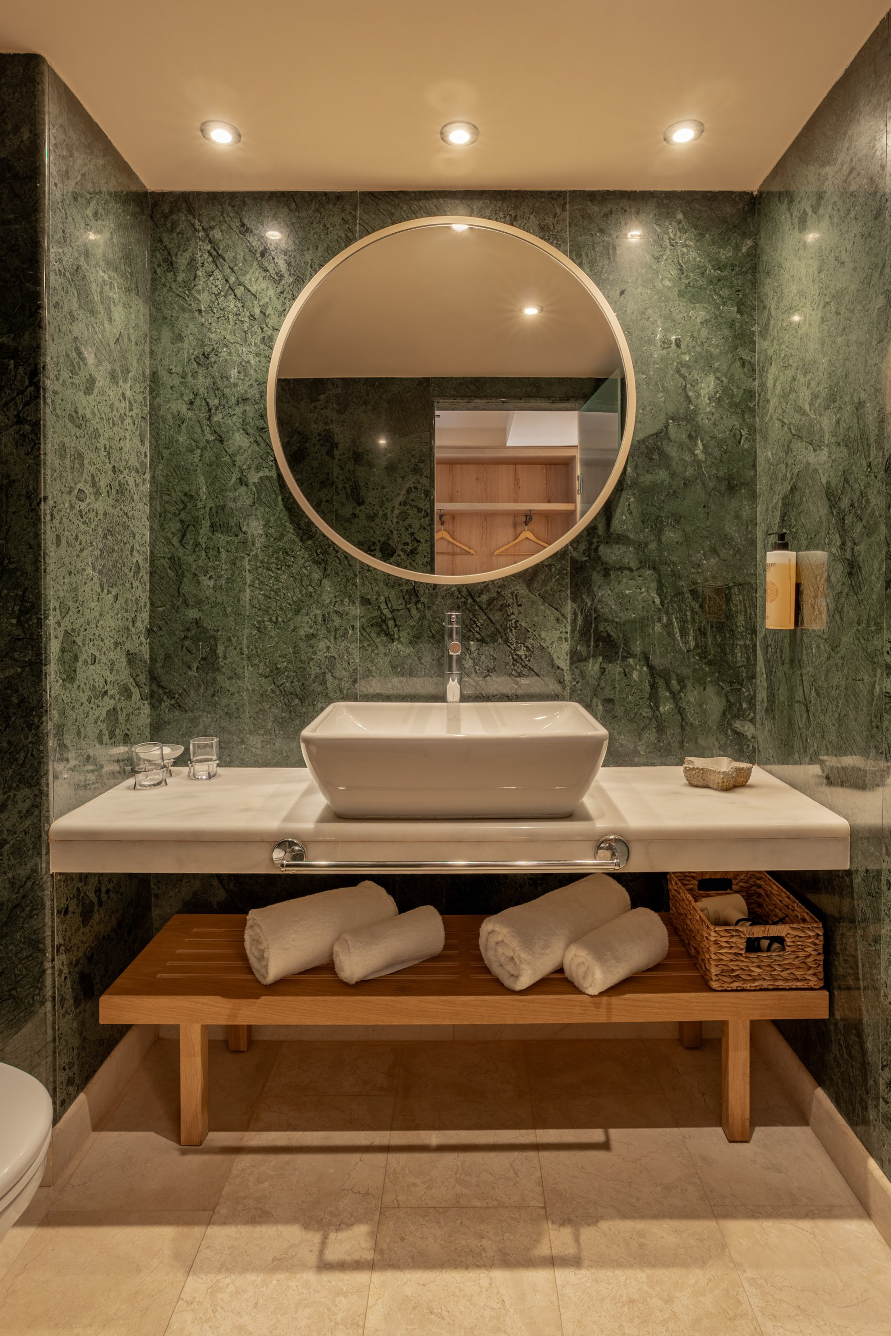 Modern bathroom with green marble, vessel sink, round mirror, and wood storage bench.